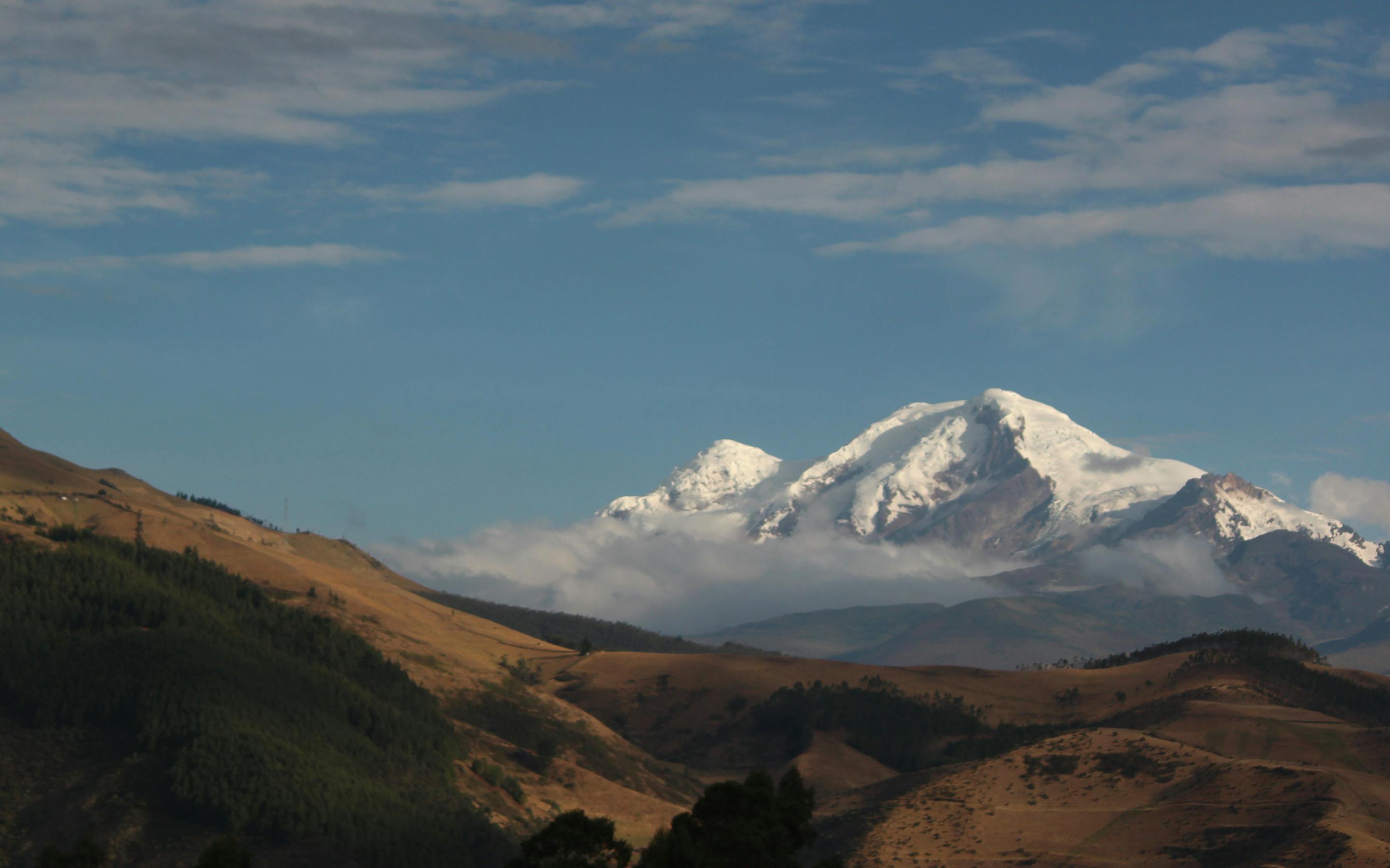 Golden hills lead to a snowcapped volcano beneath a broad blue sky, with scattered clouds over the valley.