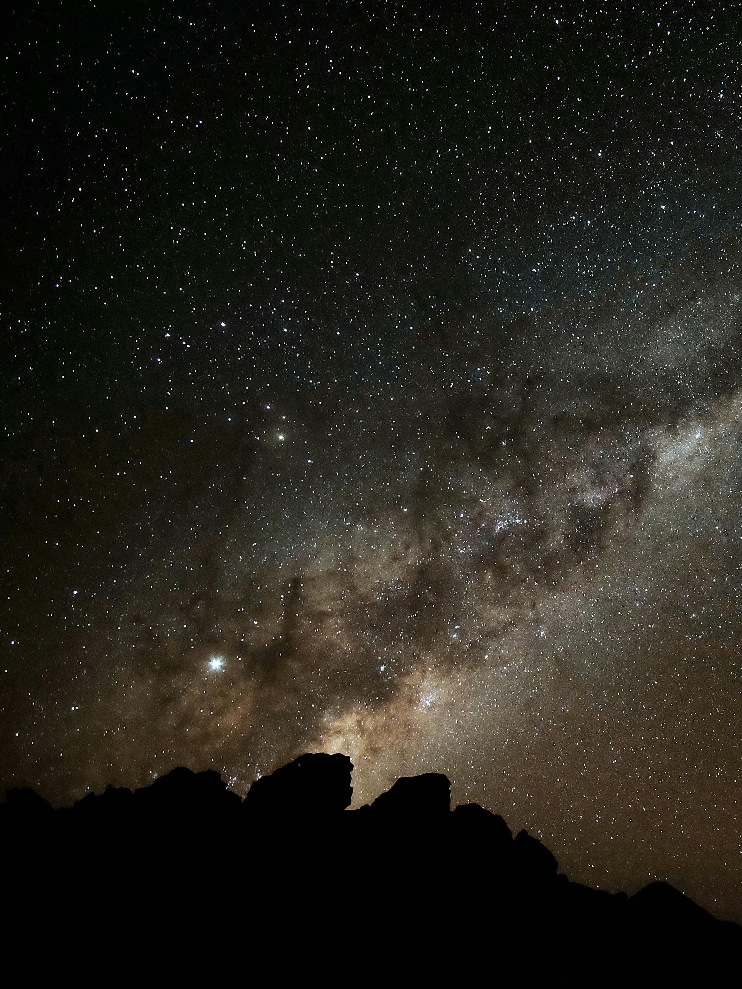 Milky Way stretches across a dark night sky above rocky silhouettes, with countless stars scattered over the horizon at night.