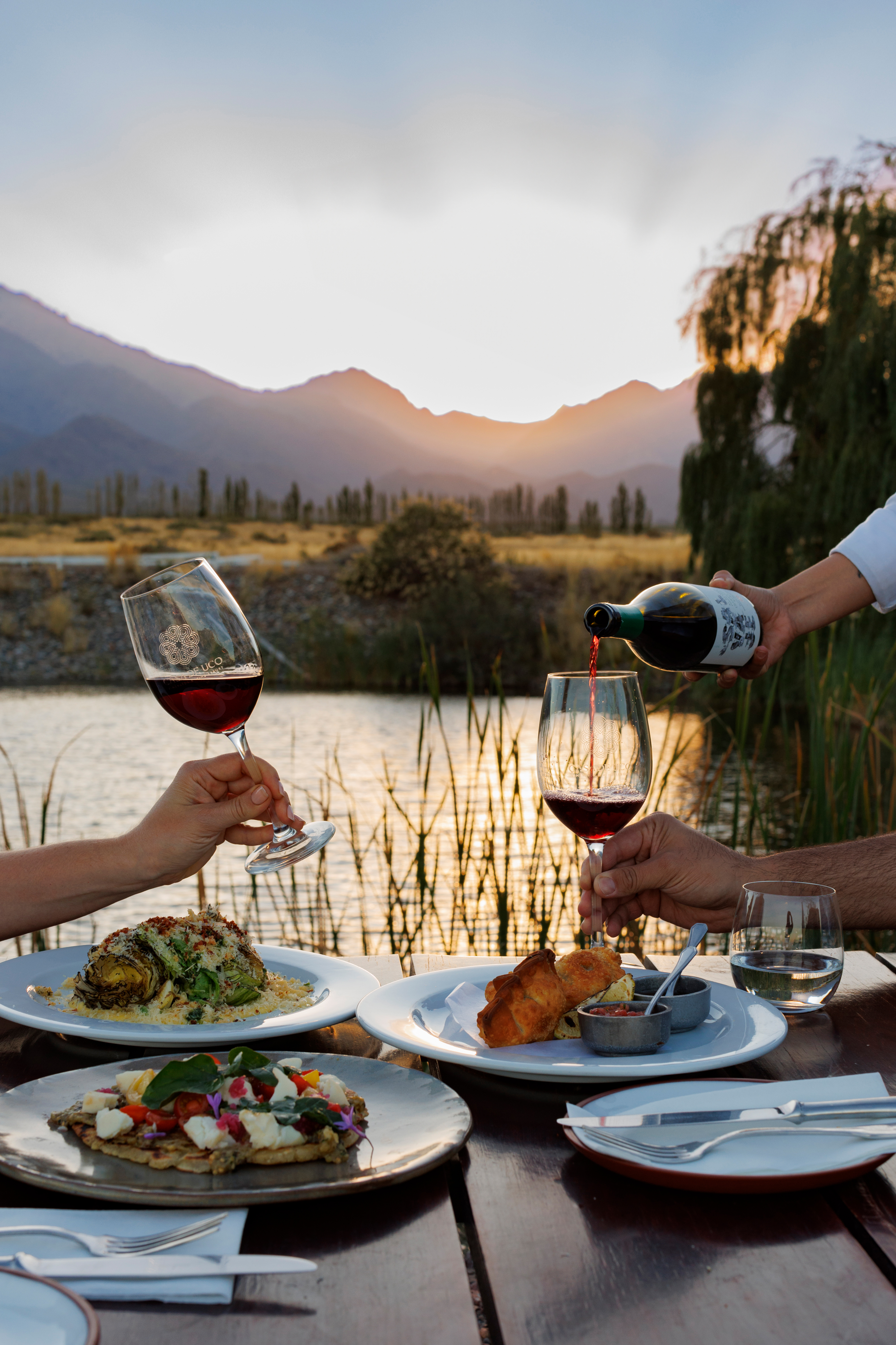 Outdoor table set with plates and wine overlooks a calm lake and mountains, with warm sunset light behind it all.