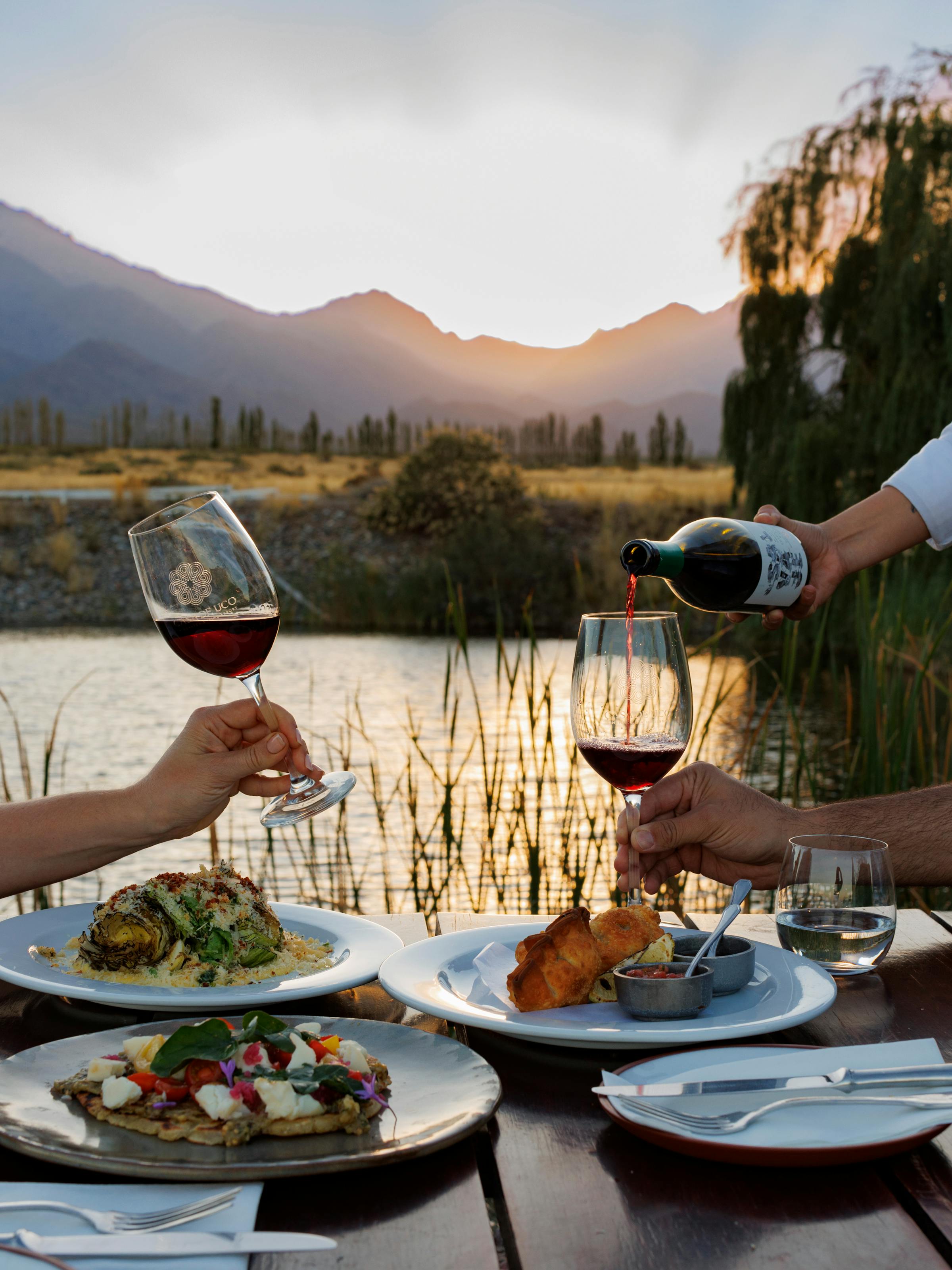 Outdoor table set with plates and wine overlooks a calm lake and mountains, with warm sunset light behind it all.