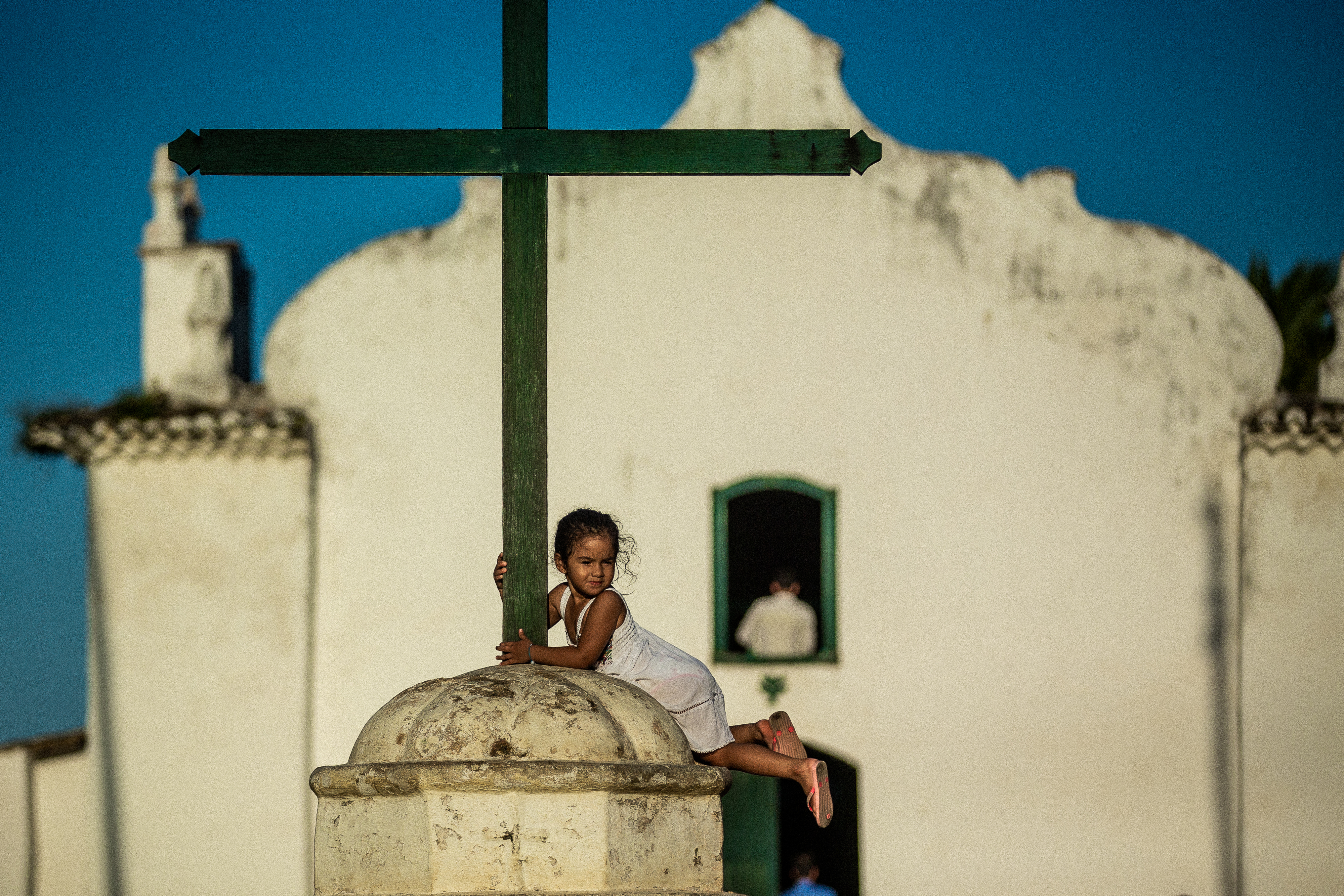 A child hugs a tall green cross atop a stone dome, with a white church facade and open doorway behind in bright sun.