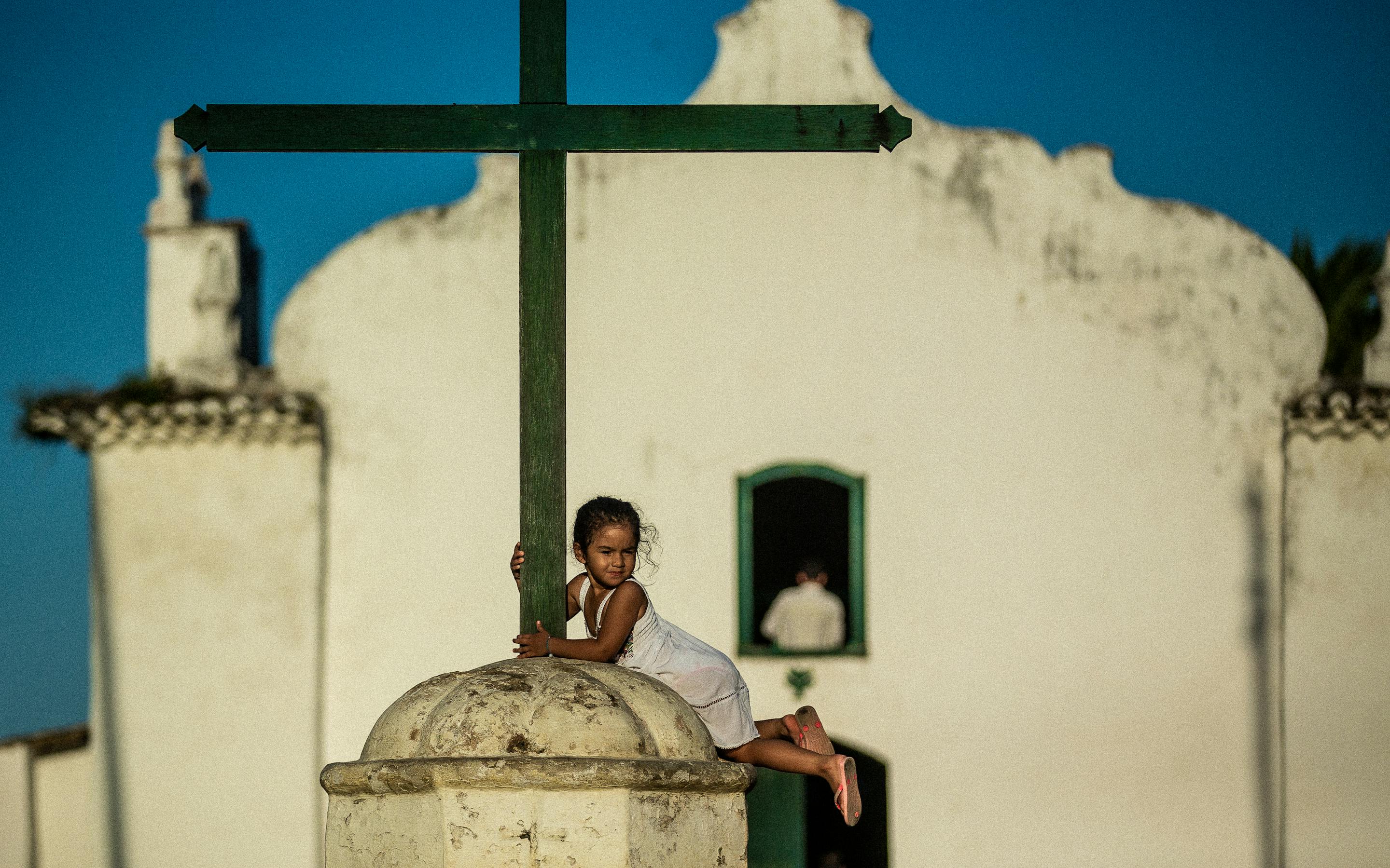 A child hugs a tall green cross atop a stone dome, with a white church facade and open doorway behind in bright sun.