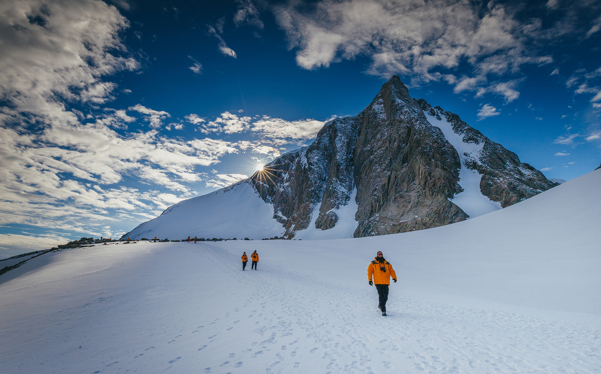 Person walks along a narrow snow ridge toward a sharp rocky peak, with dramatic clouds and bright sunlight above them.