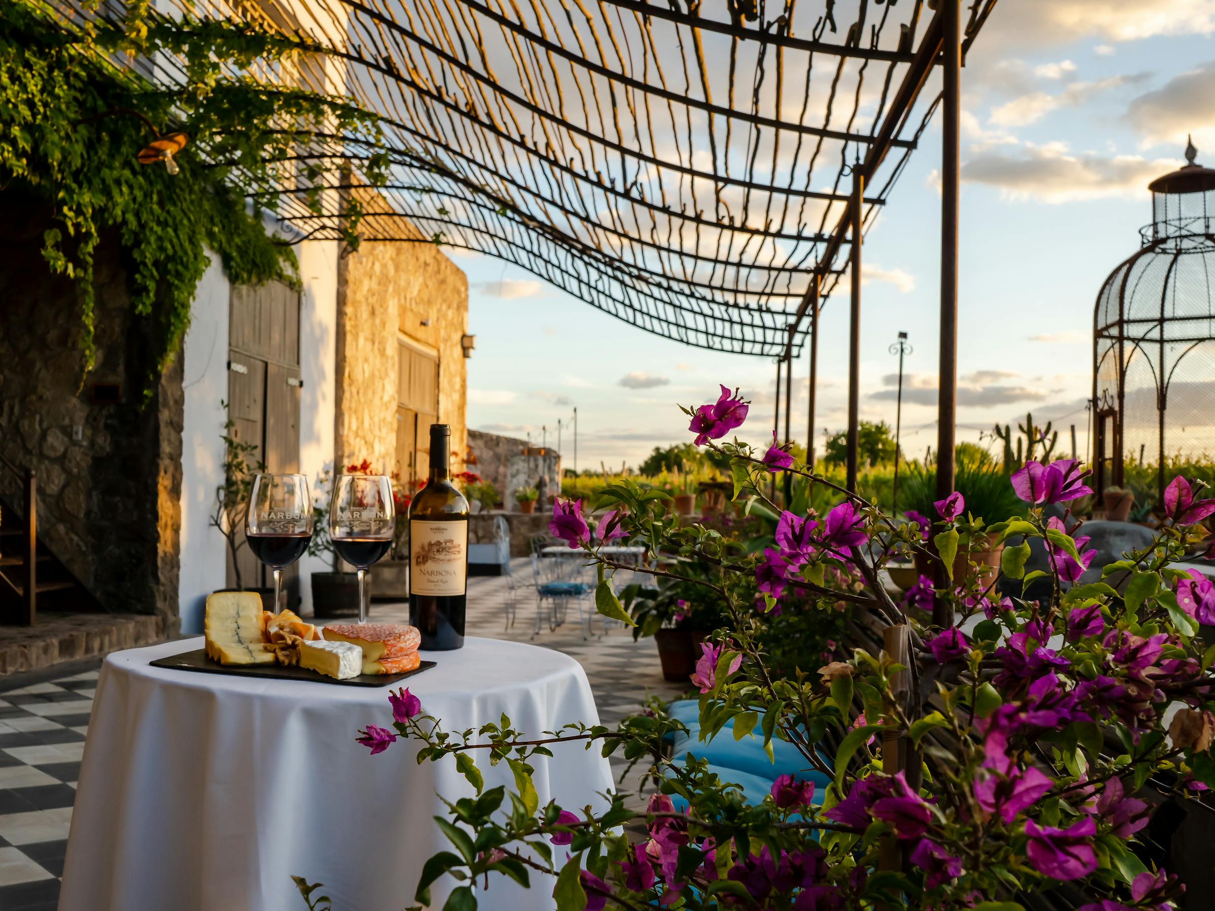 Set table with white cloth sits in a tiled courtyard beside stone arches, potted flowers, and a decorative gazebo outside.