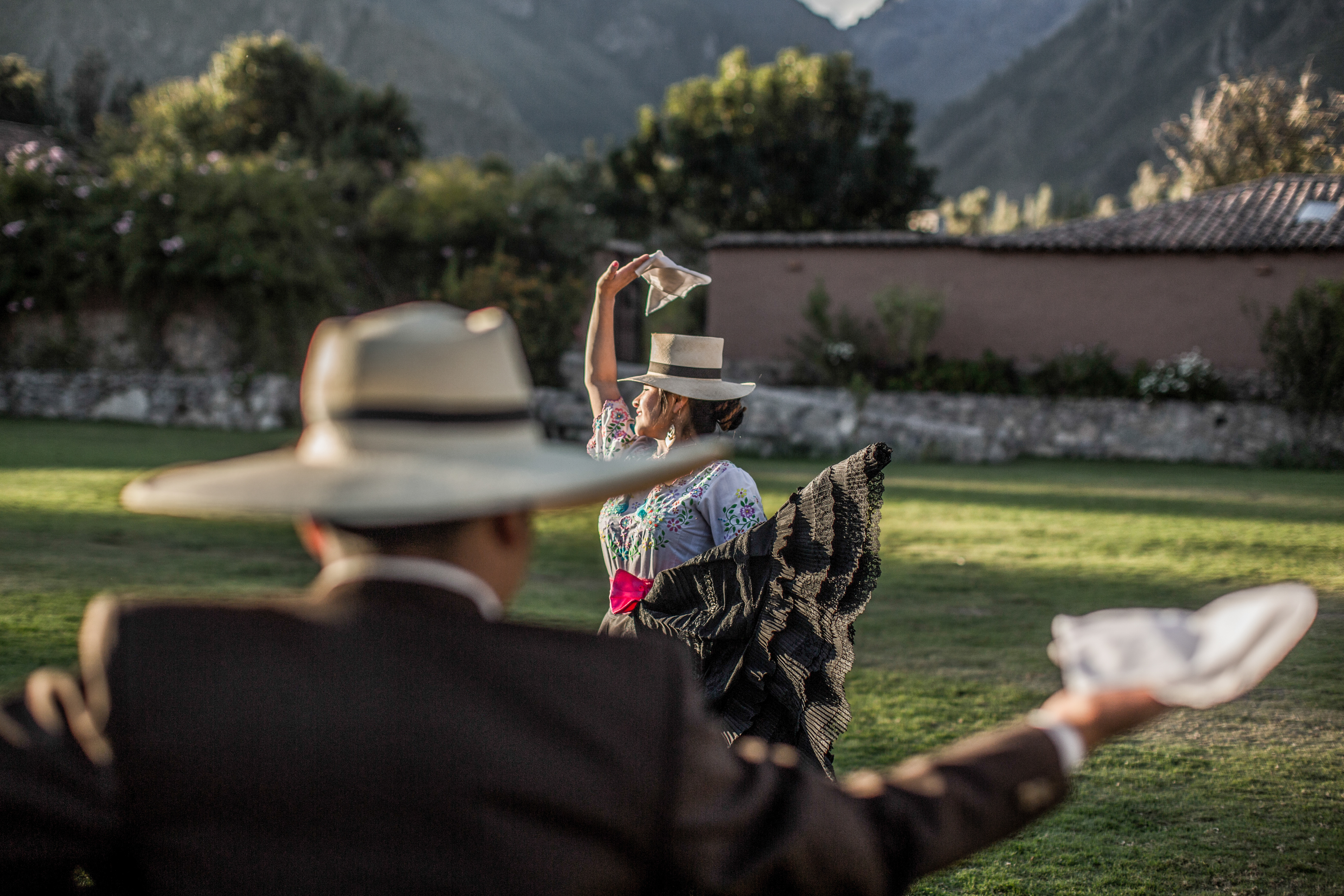 Two dancers in brimmed hats wave white handkerchiefs on a grassy field, with mountains and stone walls behind.