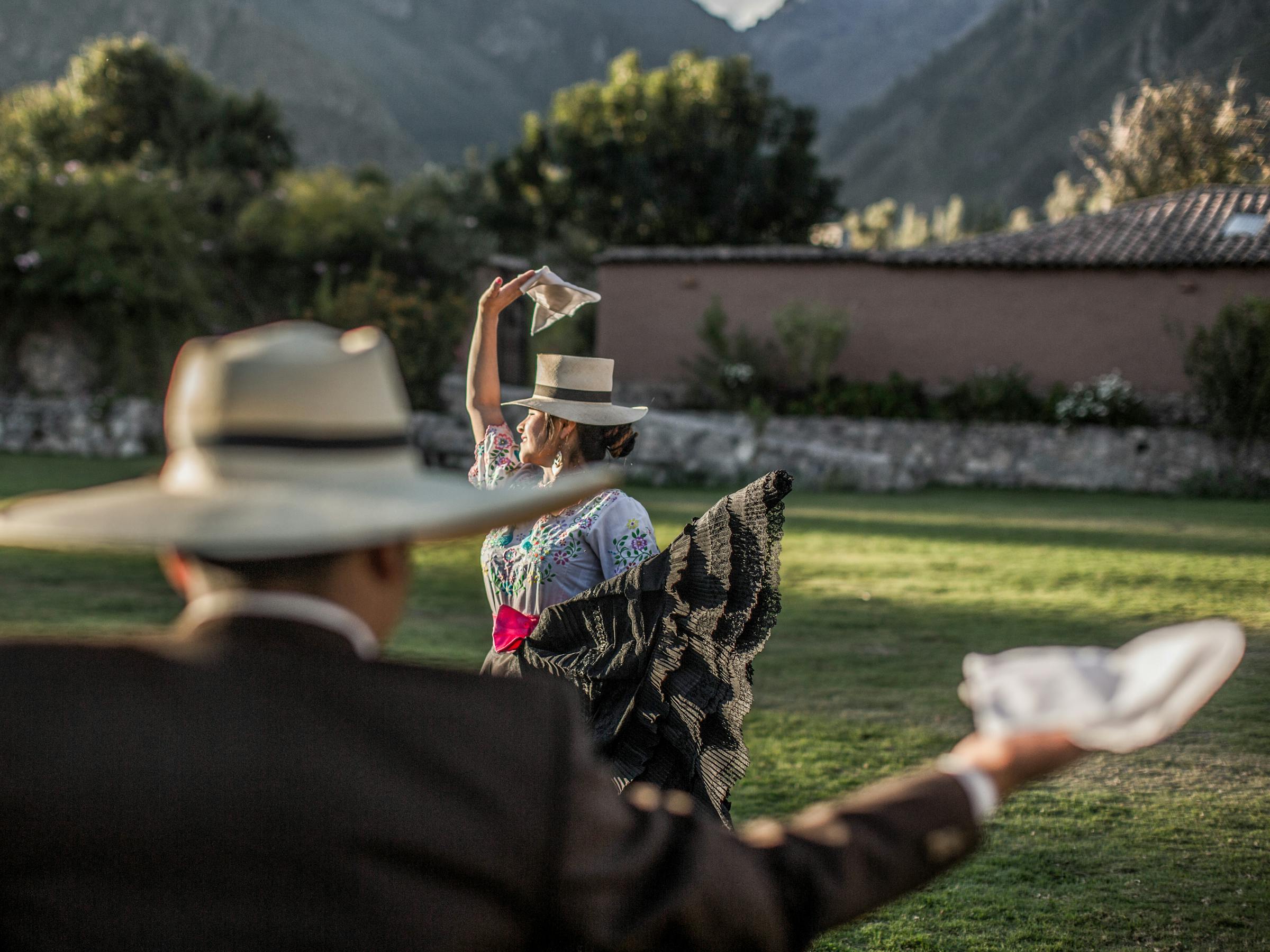 Two dancers in brimmed hats wave white handkerchiefs on a grassy field, with mountains and stone walls behind.