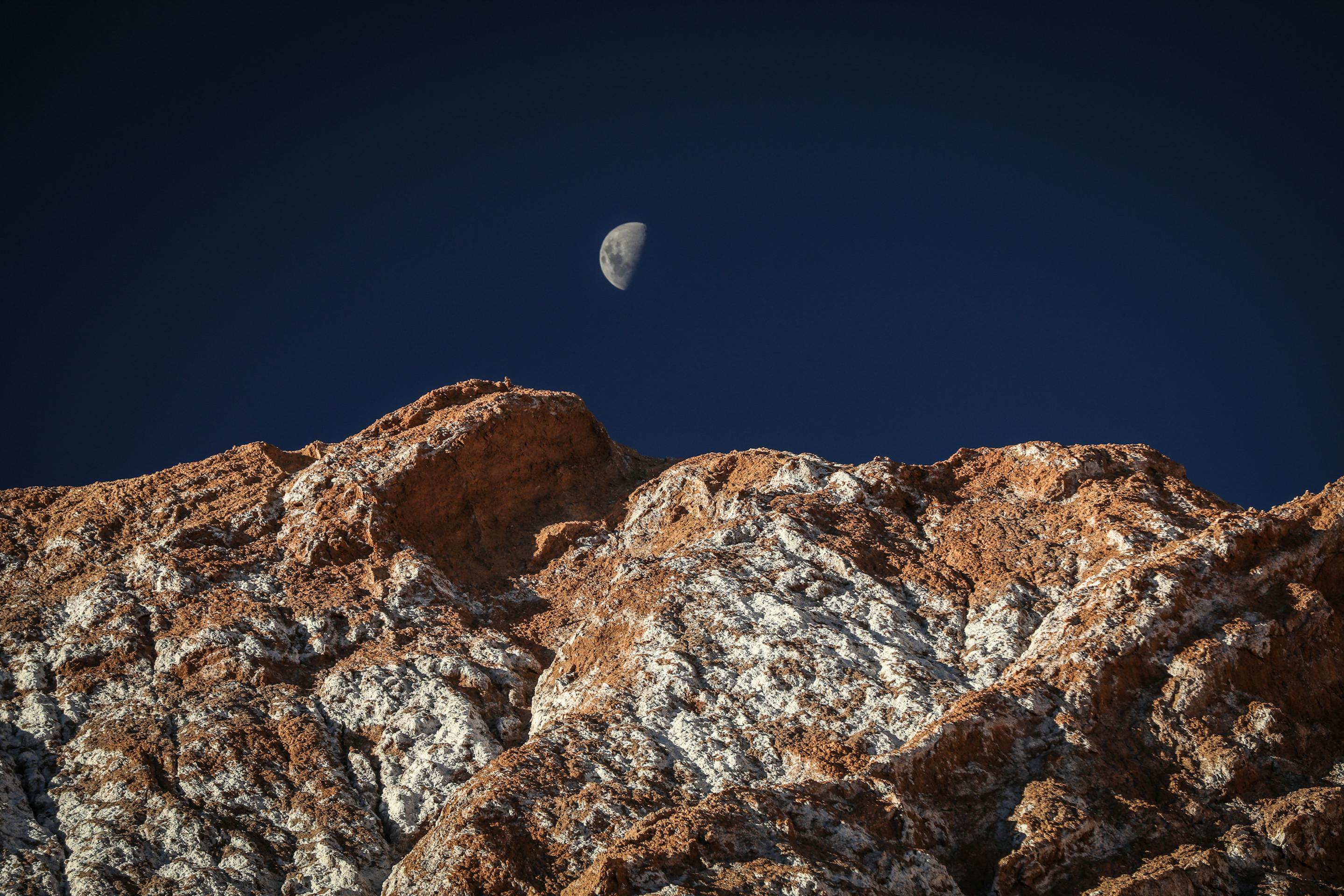 Night scene shows a bright moon above rugged rocks, with deep blue sky and faint stars over desert terrain below.