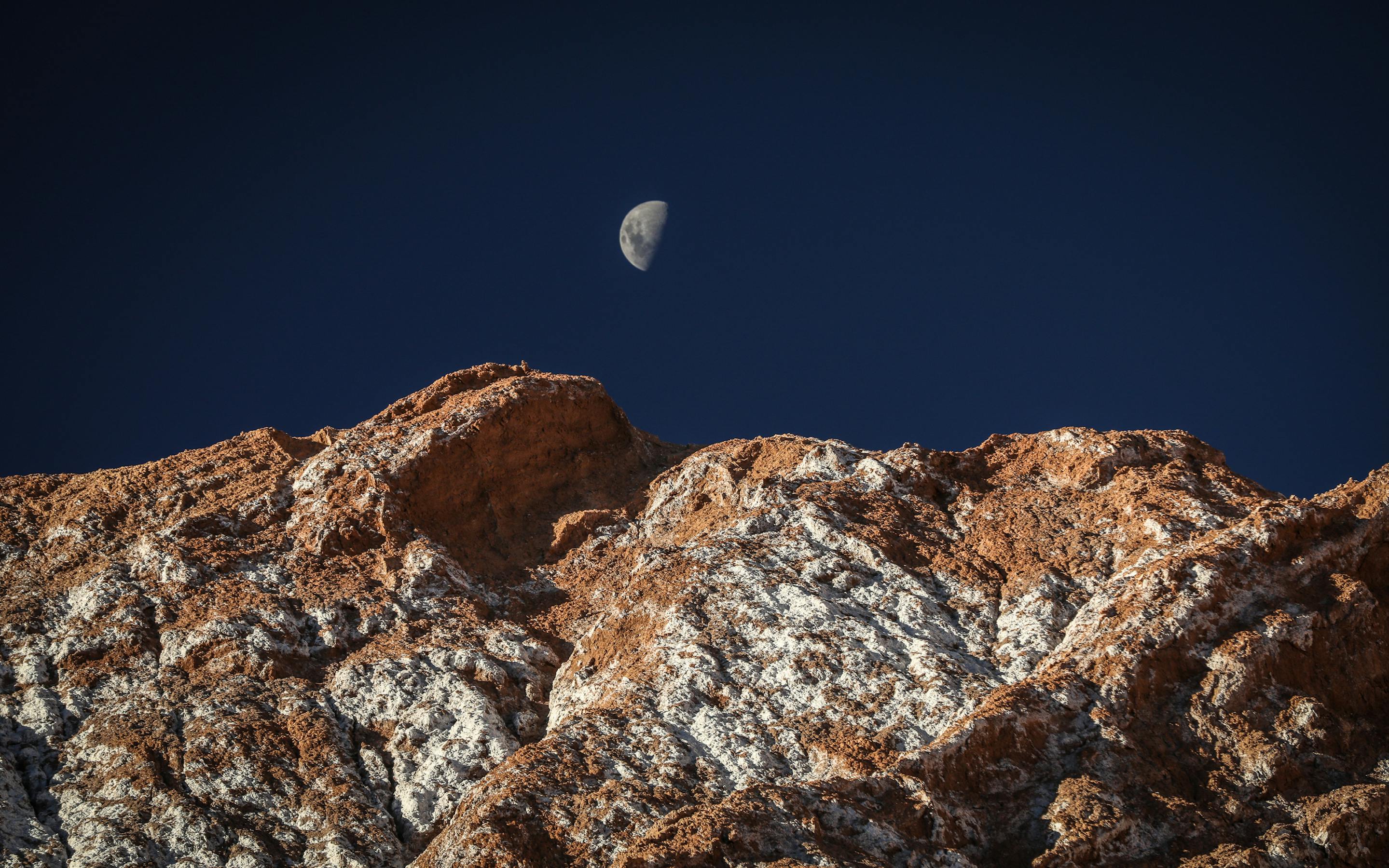 Night scene shows a bright moon above rugged rocks, with deep blue sky and faint stars over desert terrain below.