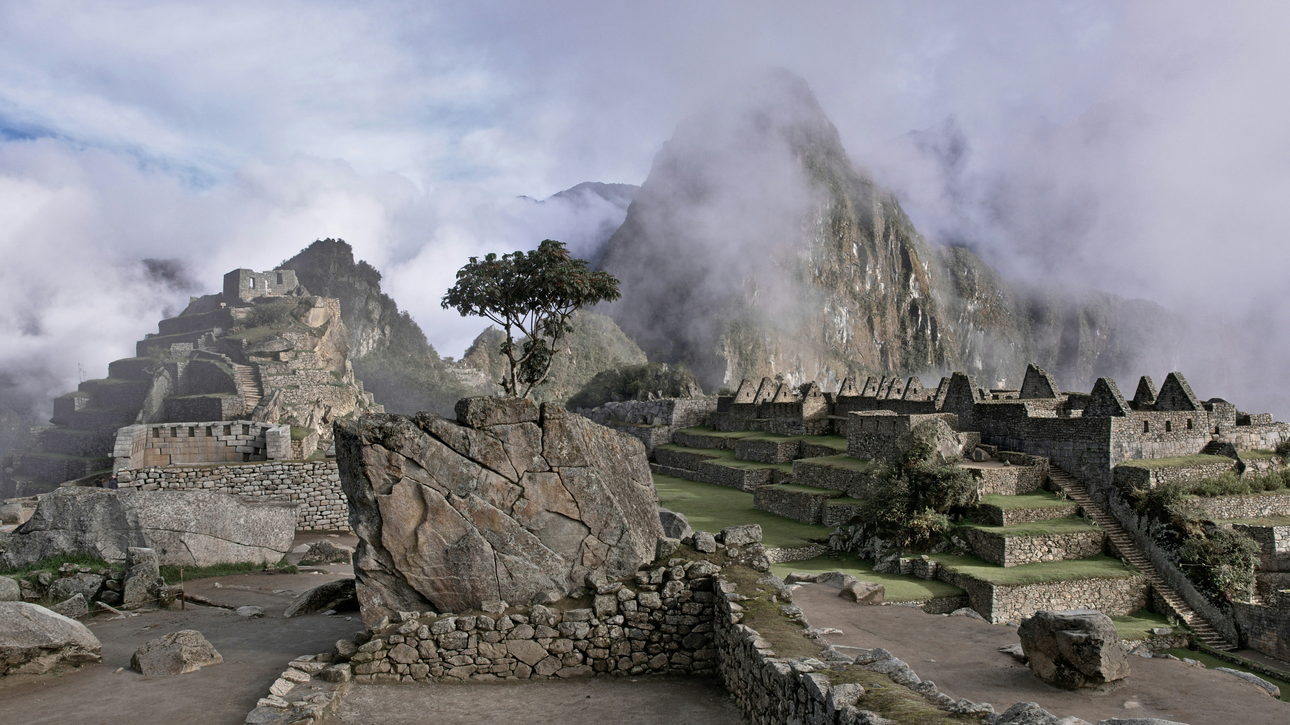 Stone ruins spread across a mountain saddle, with steep green slopes and misty clouds rolling through distant peaks near sunrise.