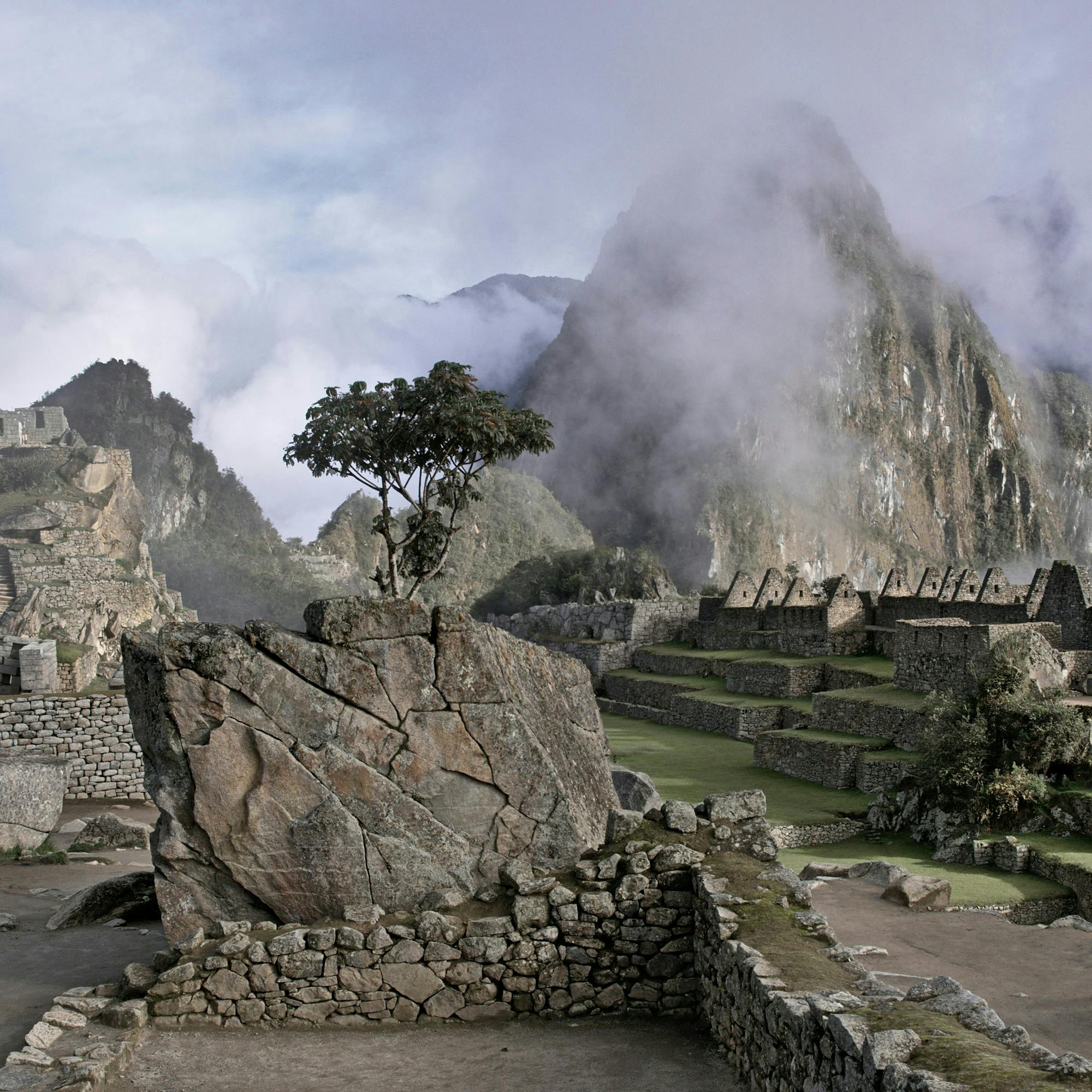 Stone ruins spread across a mountain saddle, with steep green slopes and misty clouds rolling through distant peaks near sunrise.