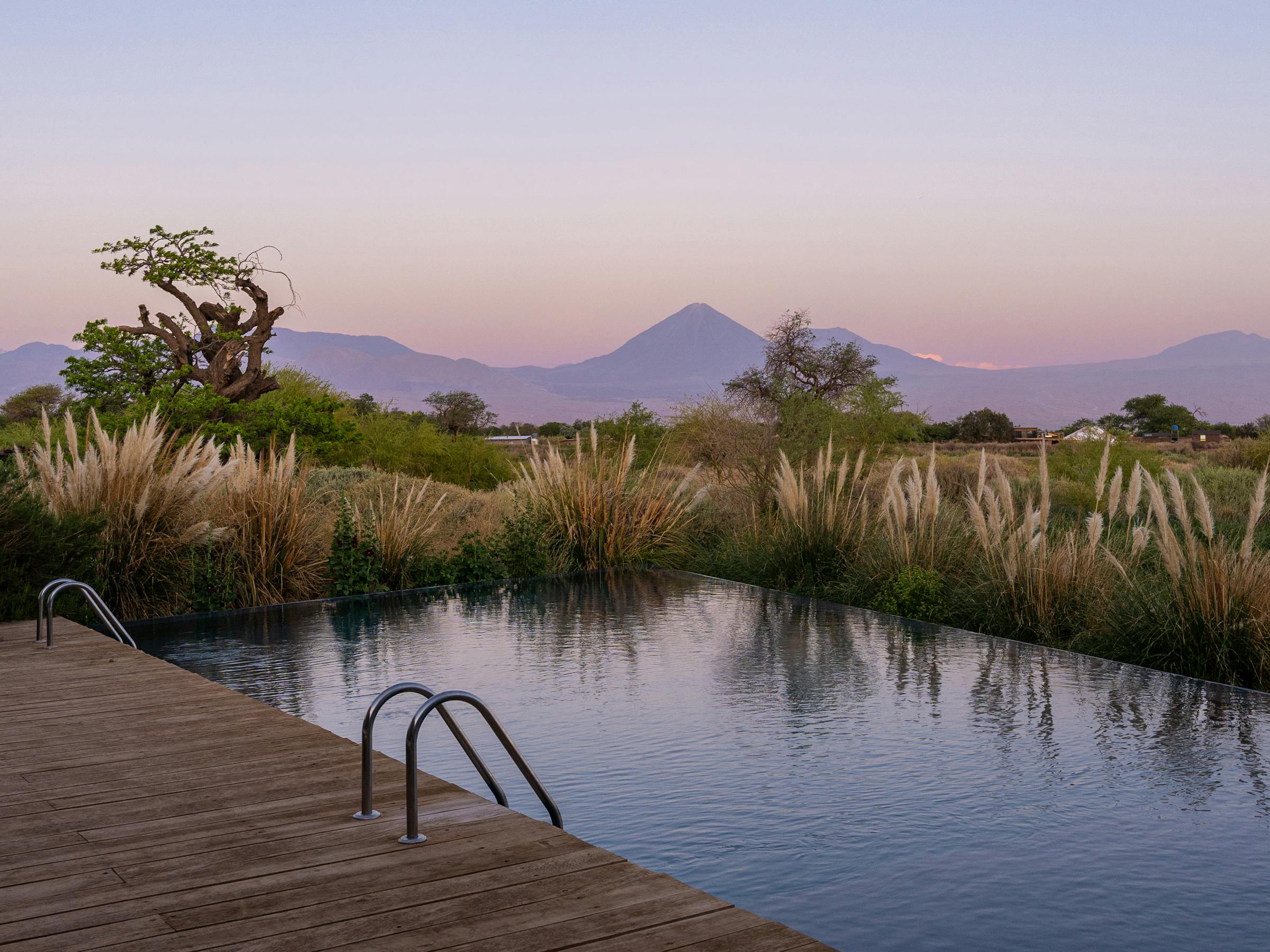 Outdoor pool reflects pink twilight sky, bordered by desert plants and distant mountains under fading evening light near the horizon.