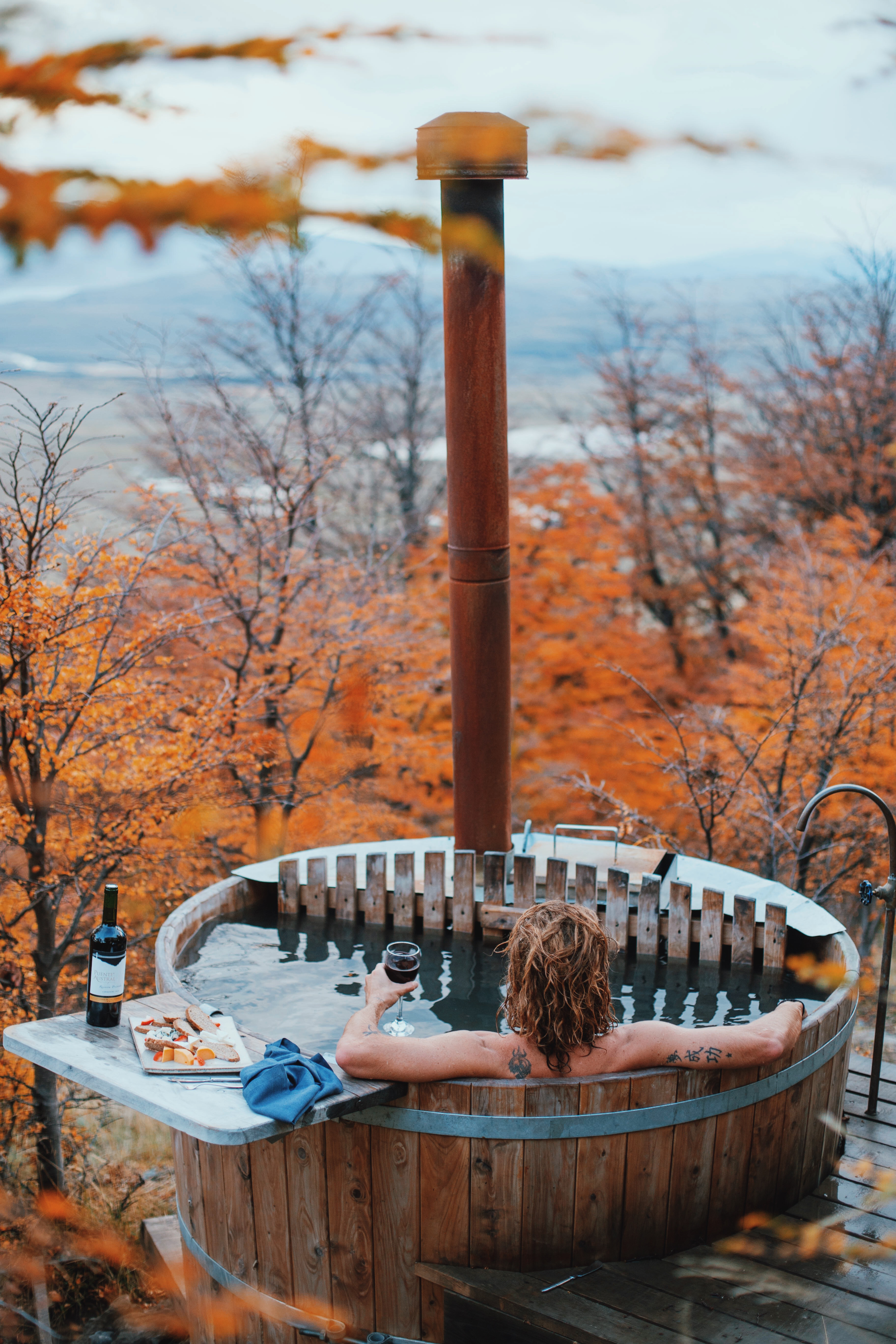 One person relaxes in a wooden barrel hot tub with a stove pipe, as autumn trees overlook calm water beyond.