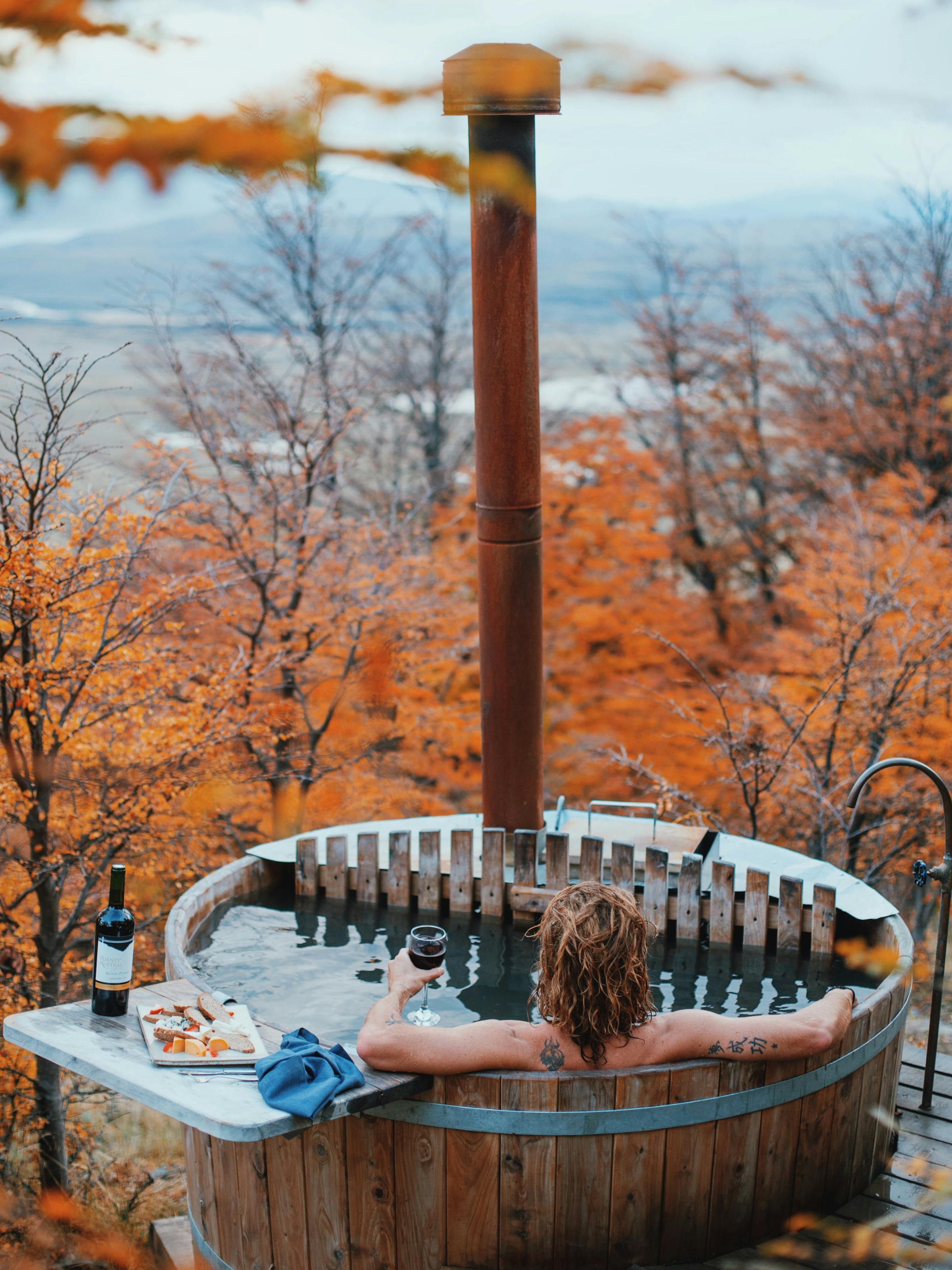 One person relaxes in a wooden barrel hot tub with a stove pipe, as autumn trees overlook calm water beyond.