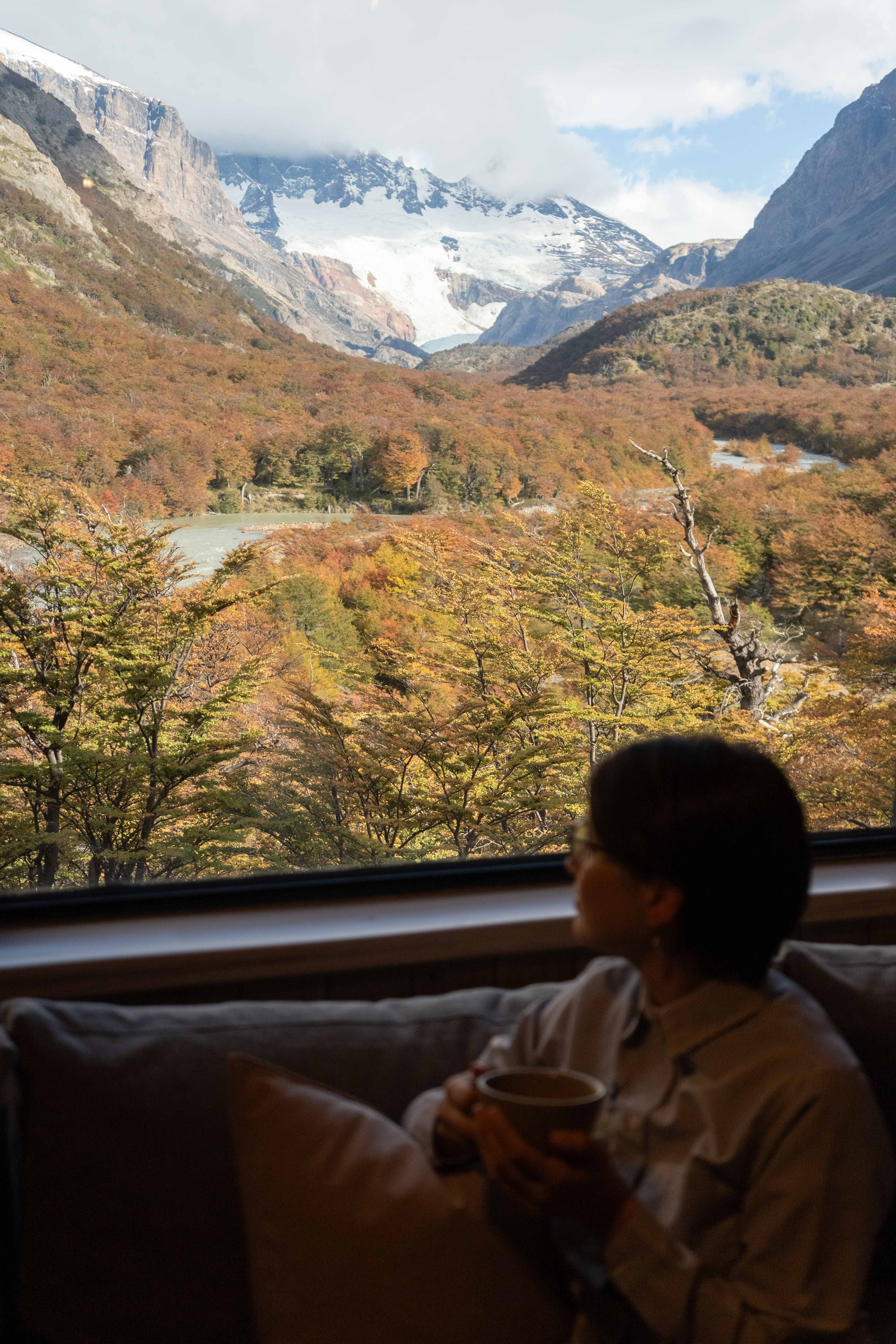 Person sits by a window in a cozy room, looking out at a river valley and rugged mountains beyond.