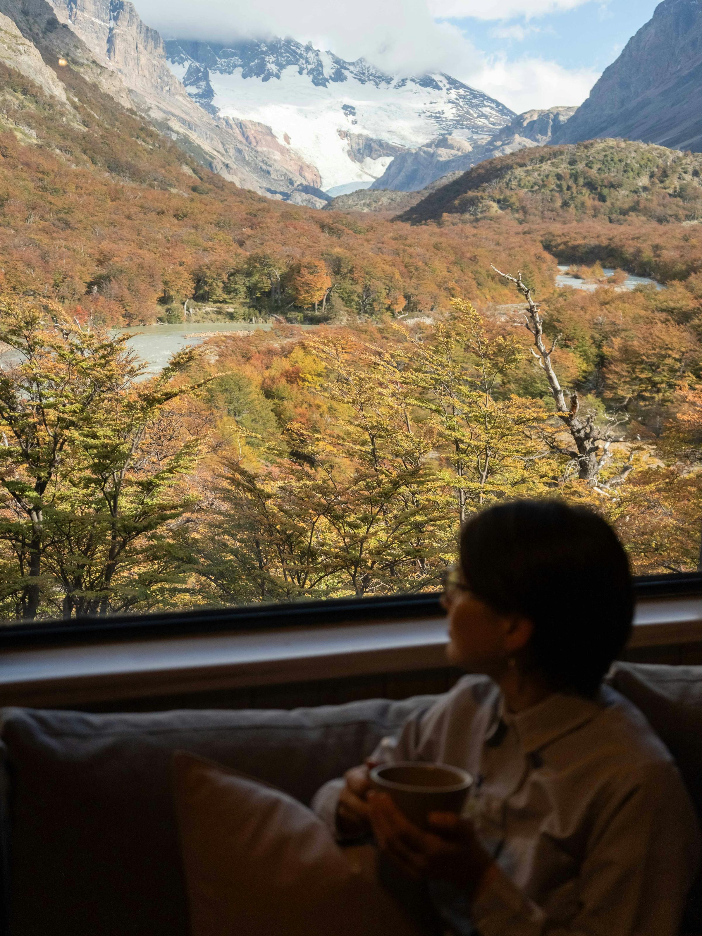 Person sits by a window in a cozy room, looking out at a river valley and rugged mountains beyond.