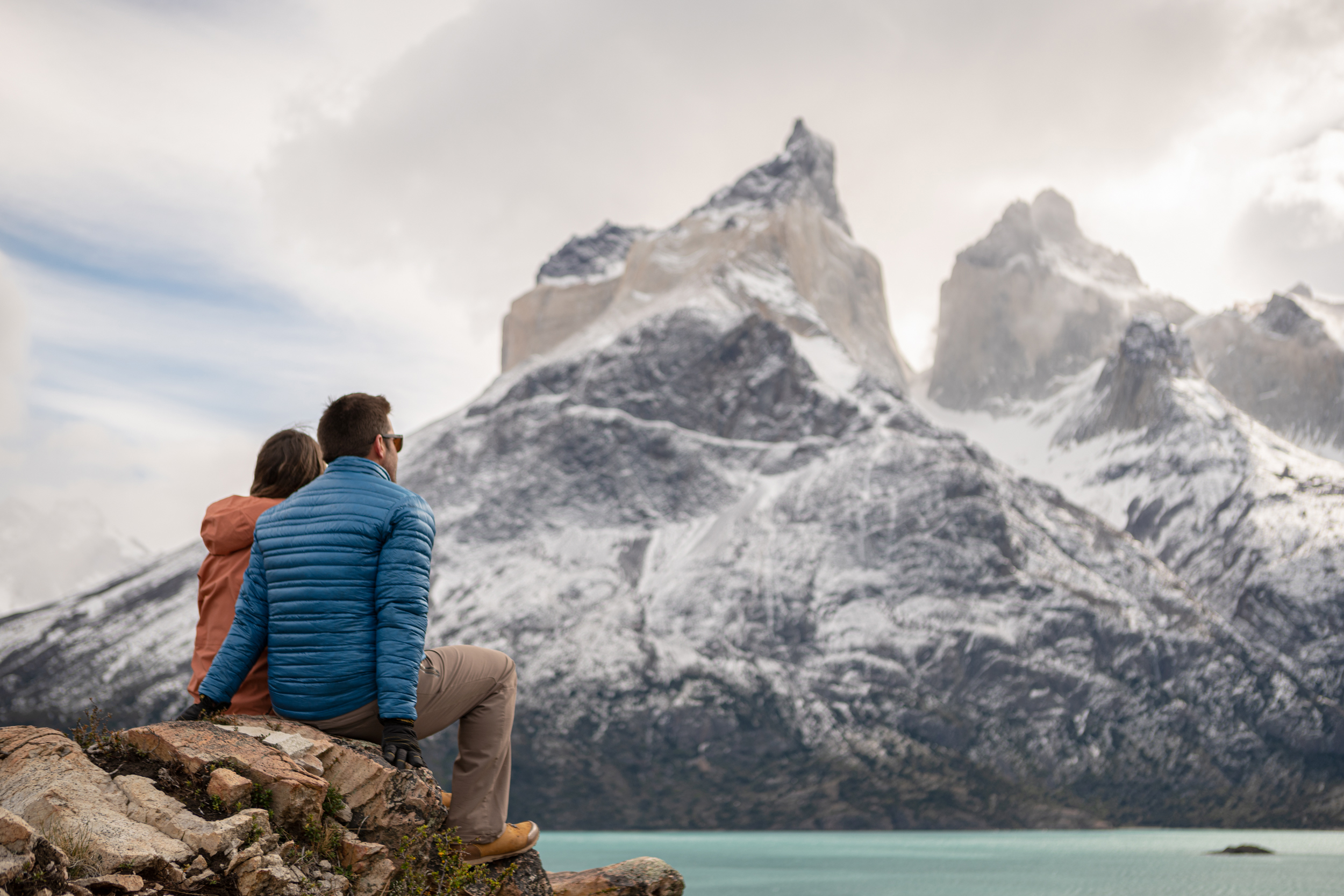 Two people sit on rocks by a teal lake, watching towering peaks rising through mist and low clouds ahead quietly.