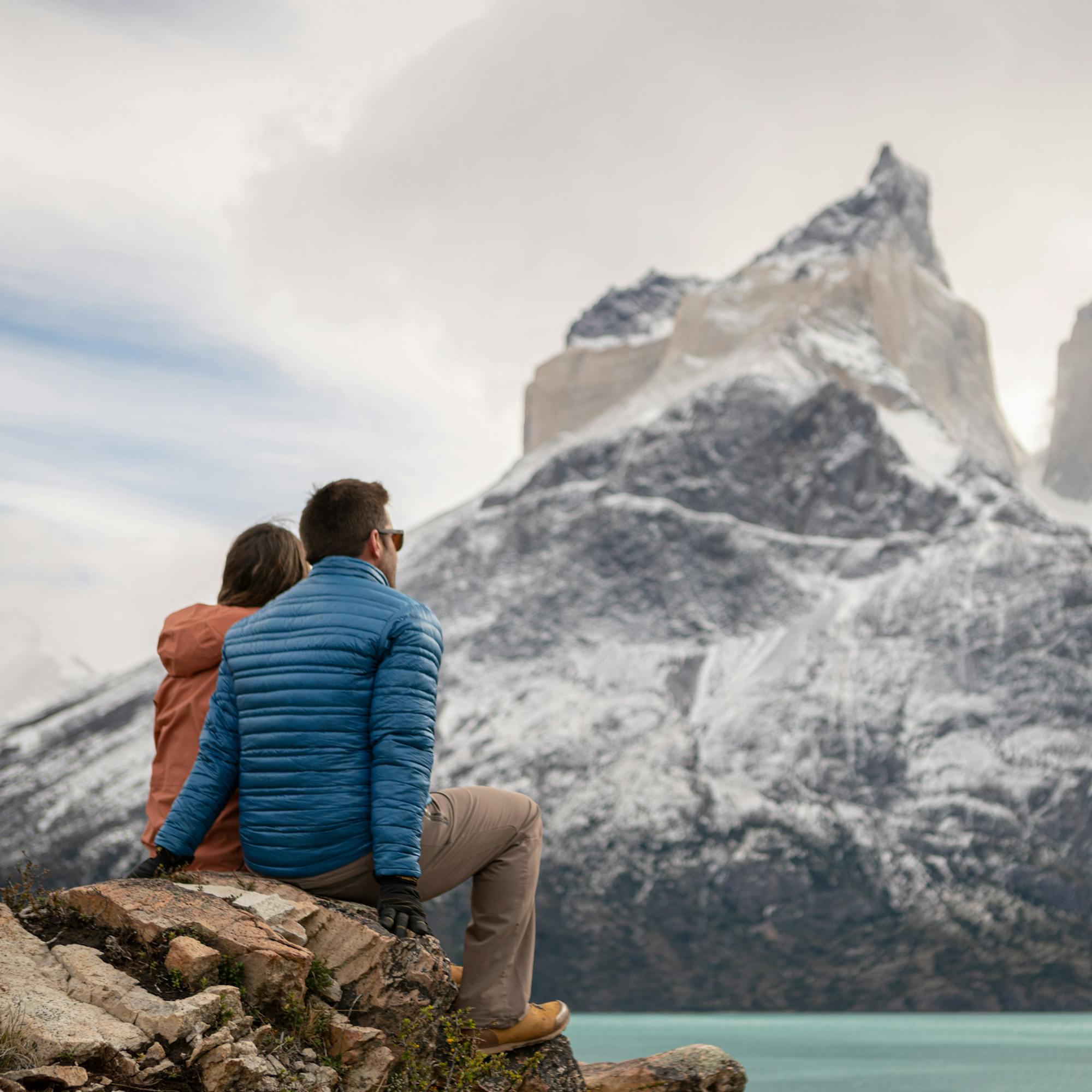 Two people sit on rocks by a teal lake, watching towering peaks rising through mist and low clouds ahead quietly.