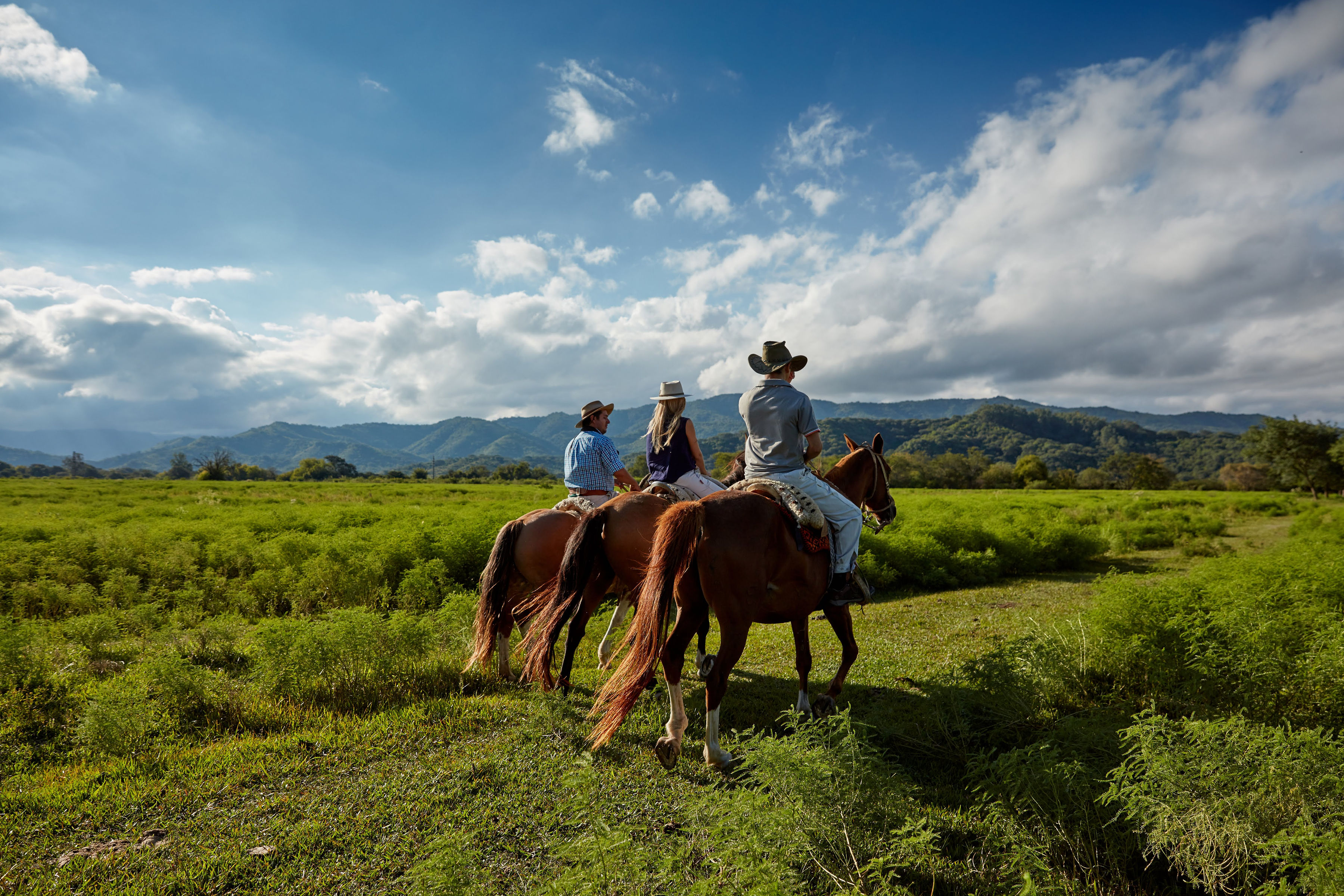 Riders on horseback cross green pasture under big skies, with low mountains and clouds forming the backdrop behind them.