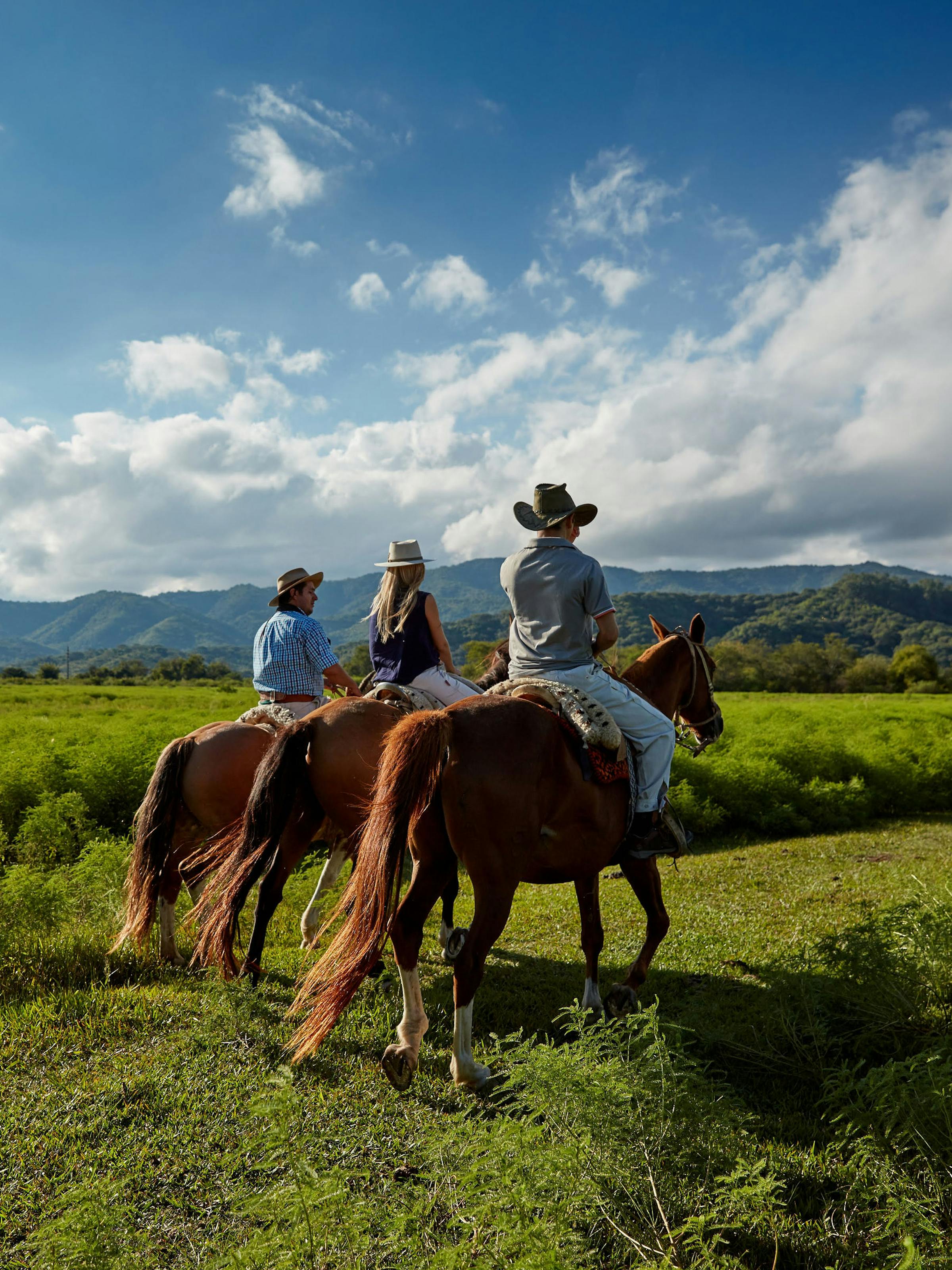 Riders on horseback cross green pasture under big skies, with low mountains and clouds forming the backdrop behind them.