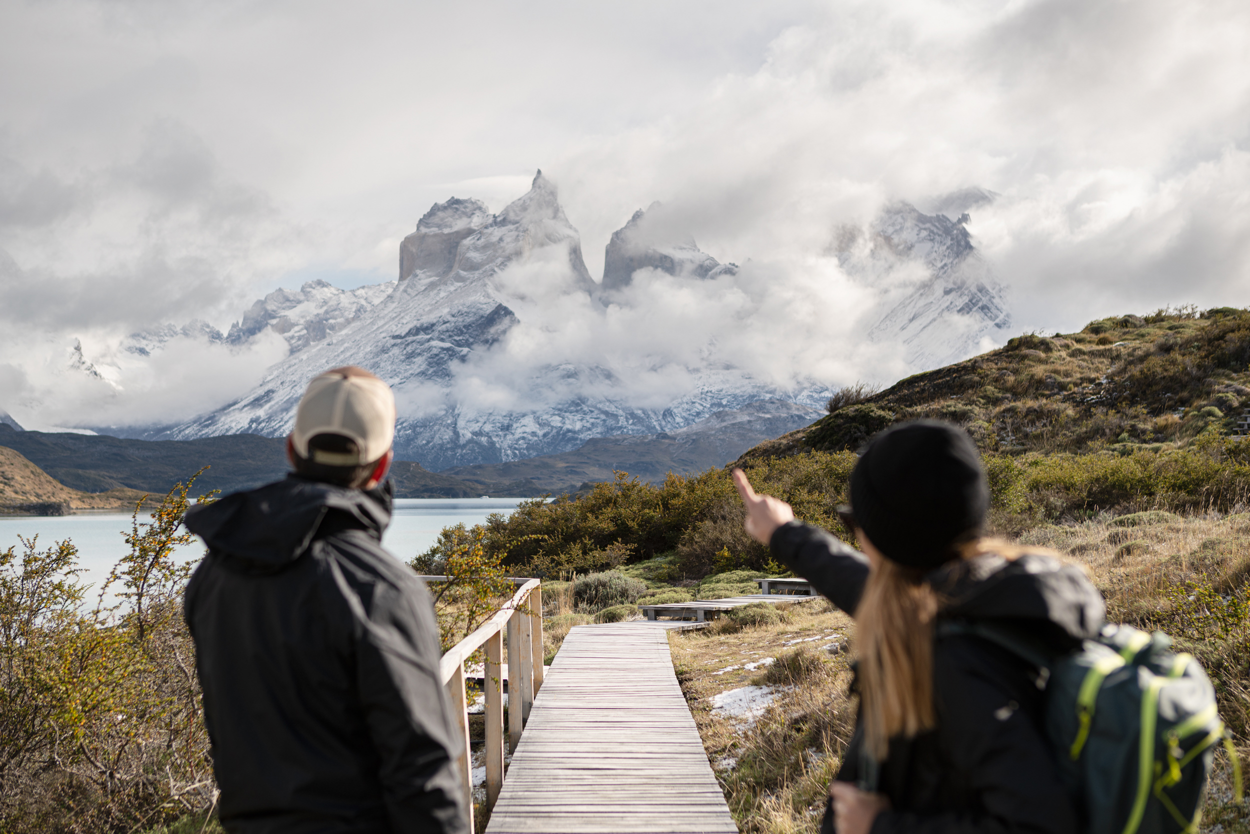 Two hikers pause on a wooden boardwalk, pointing toward snowy mountains and low clouds in a rugged valley ahead.