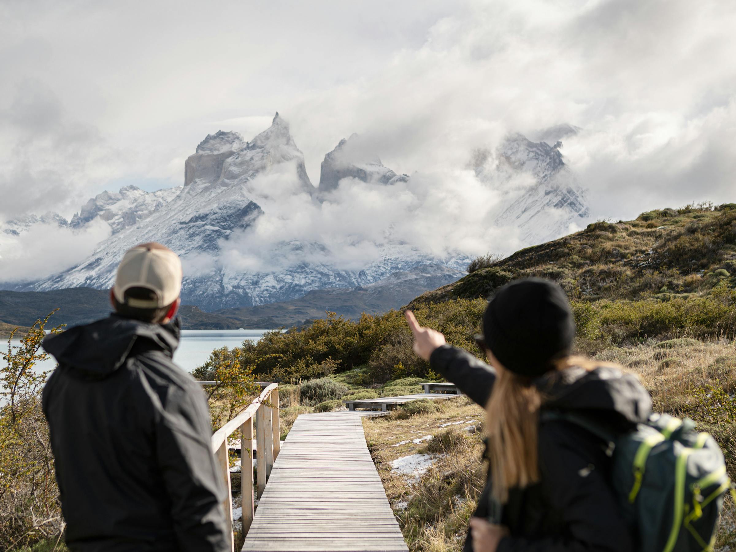 Two hikers pause on a wooden boardwalk, pointing toward snowy mountains and low clouds in a rugged valley ahead.