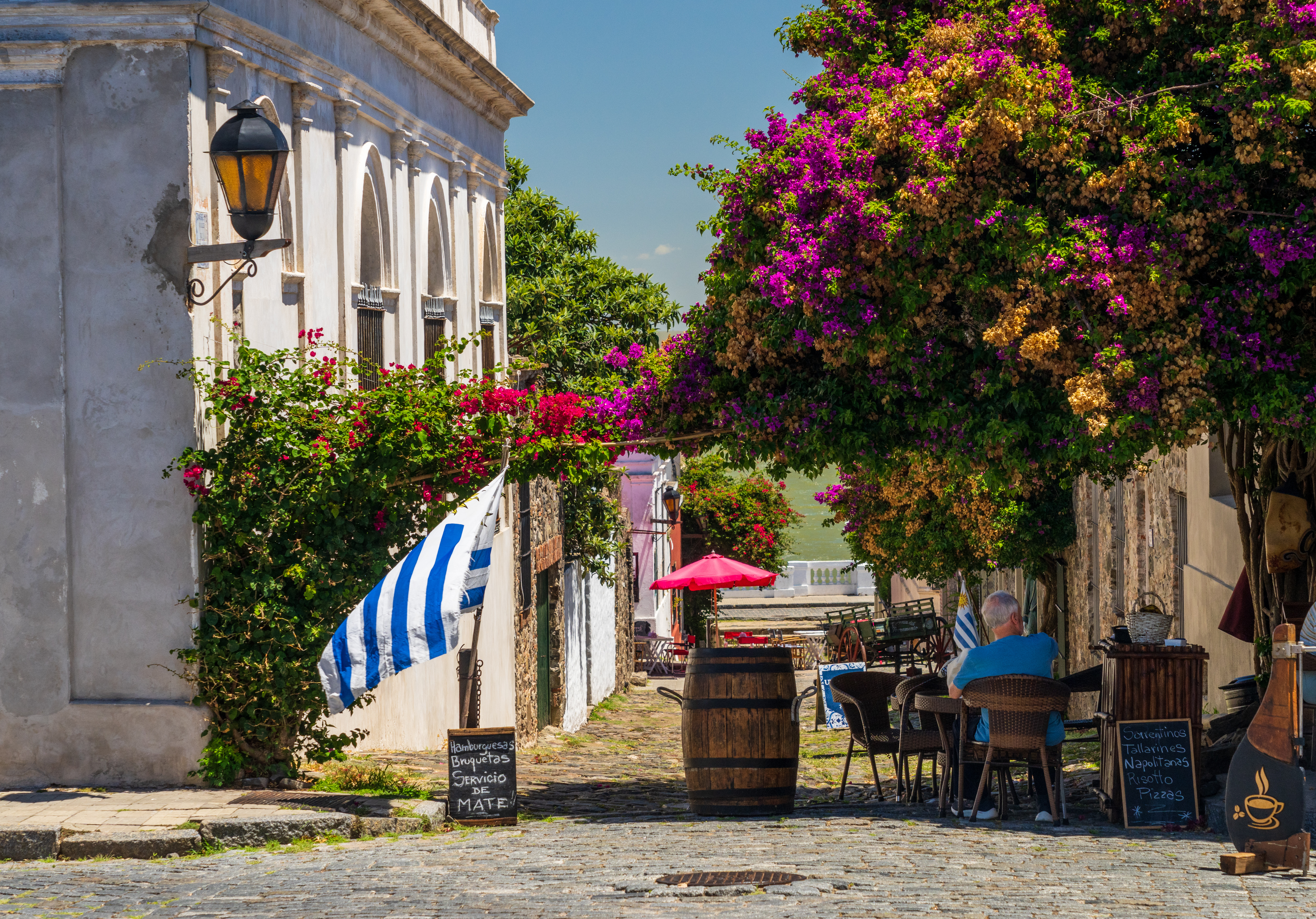 A cobblestone street cafe sits under vivid bougainvillea, with a striped flag, barrels, and one diner at a table.