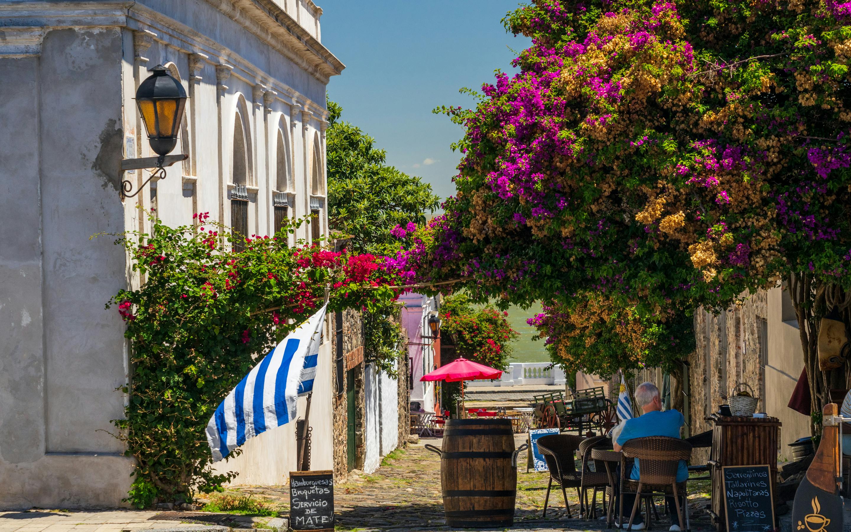 A cobblestone street cafe sits under vivid bougainvillea, with a striped flag, barrels, and one diner at a table.