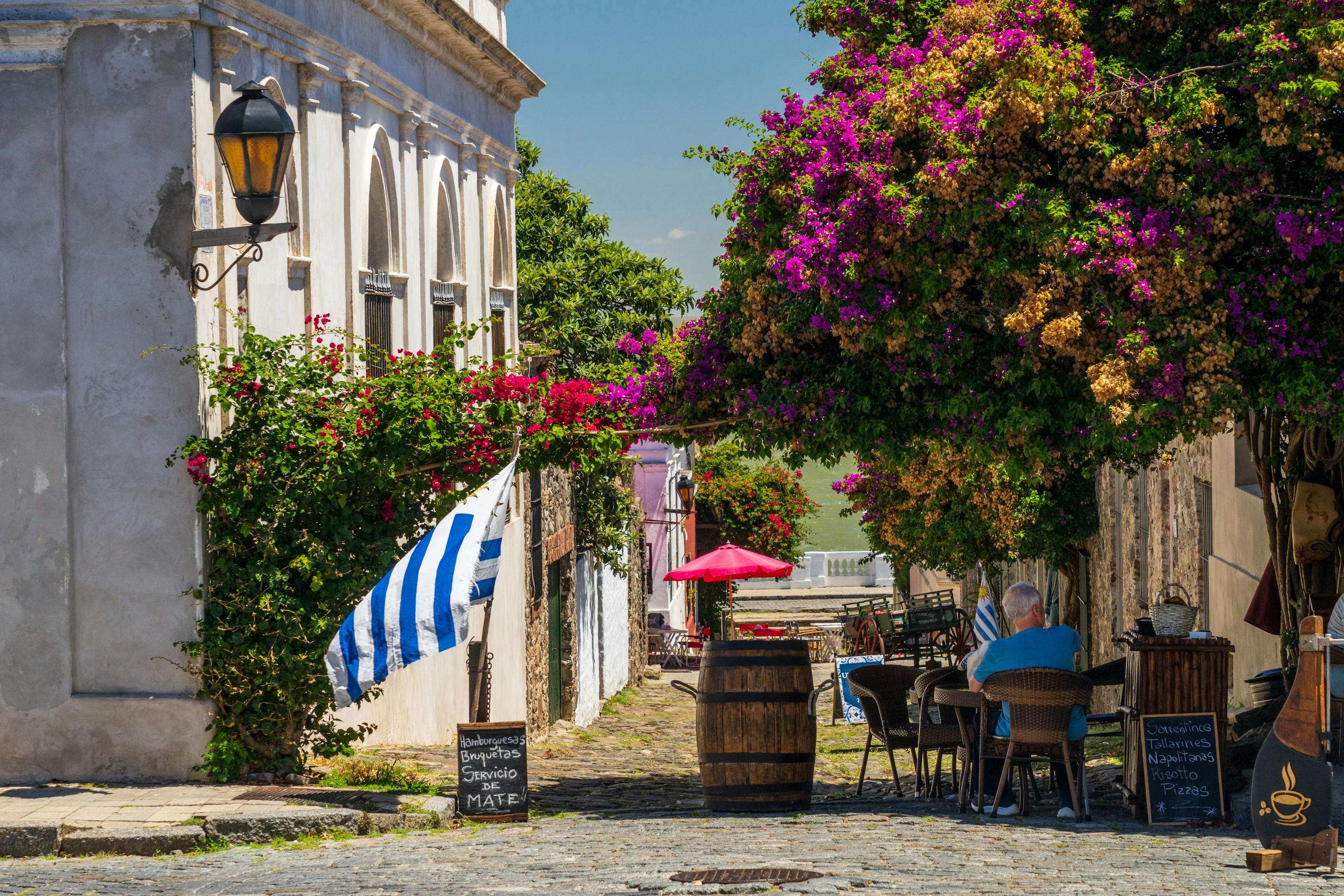 A cobblestone street cafe sits under vivid bougainvillea, with a striped flag, barrels, and one diner at a table.