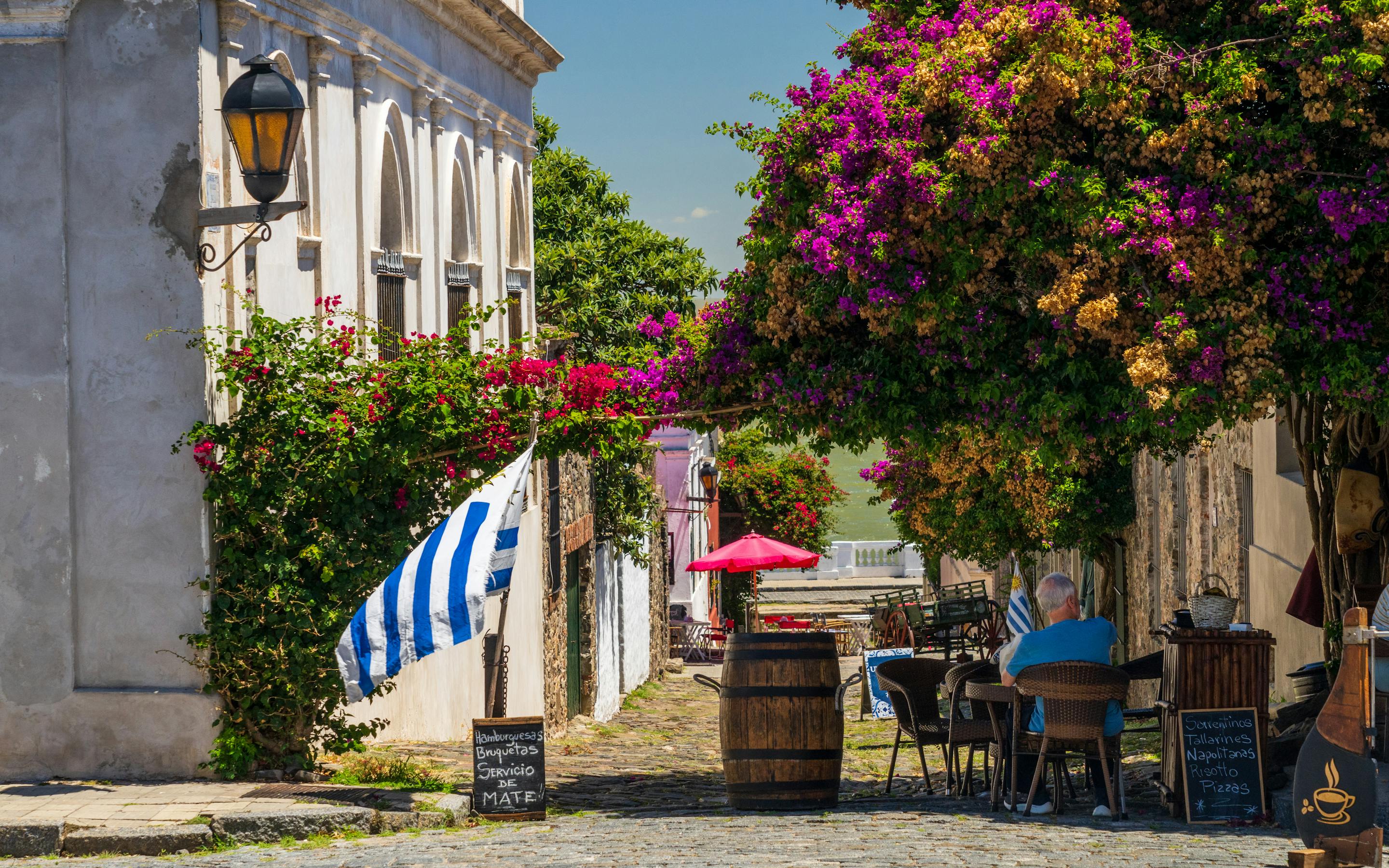 A cobblestone street cafe sits under vivid bougainvillea, with a striped flag, barrels, and one diner at a table.