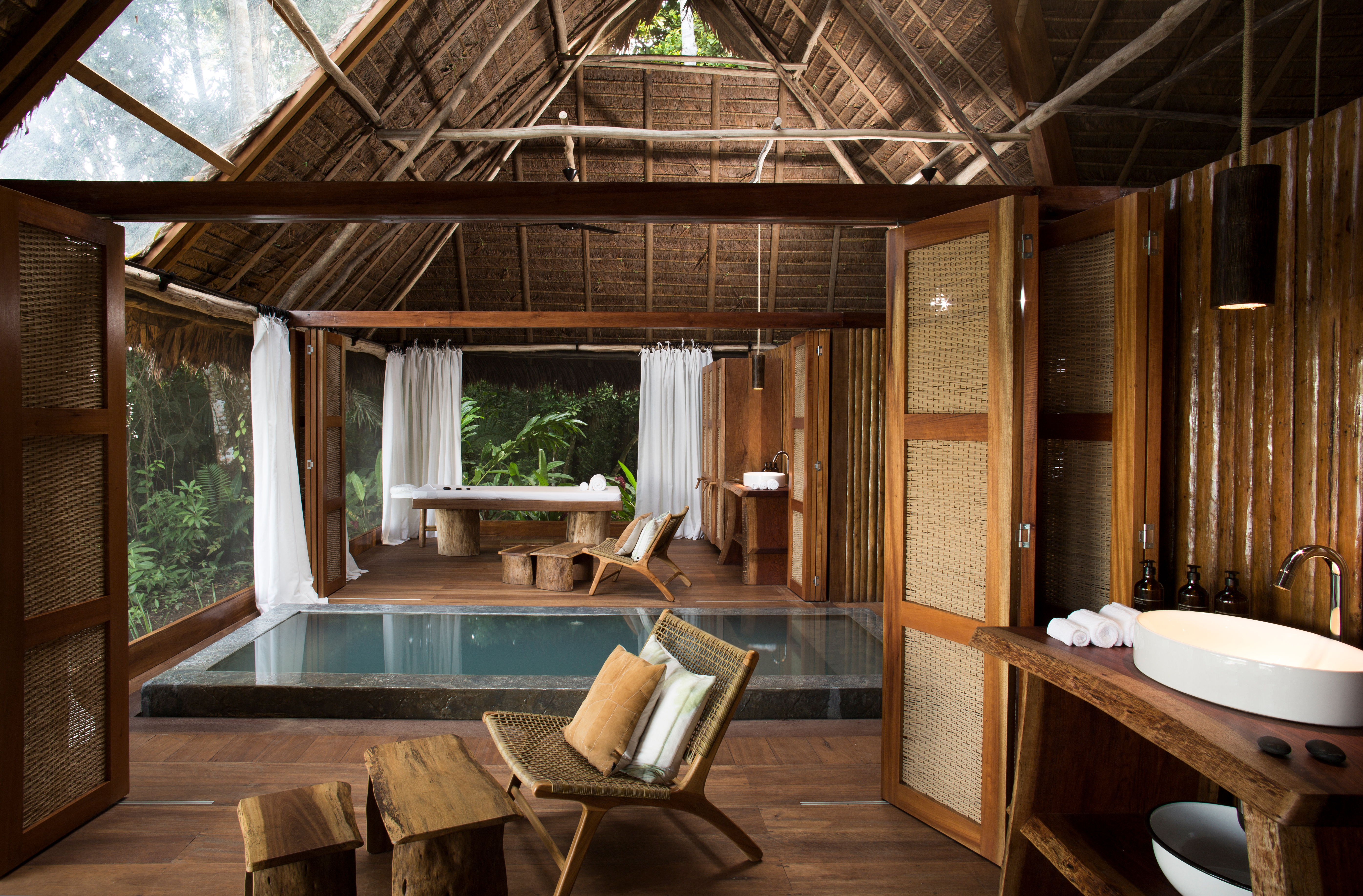 Rustic lodge room with wooden beams, open doors, and a large soaking tub looking out to green forest views outside.