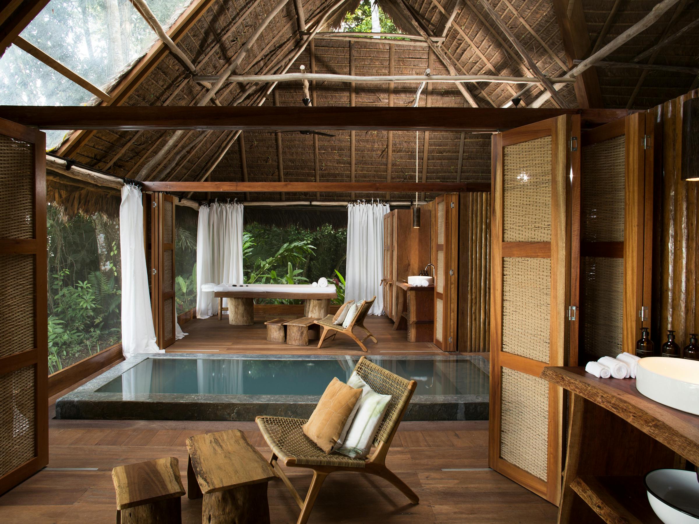 Rustic lodge room with wooden beams, open doors, and a large soaking tub looking out to green forest views outside.