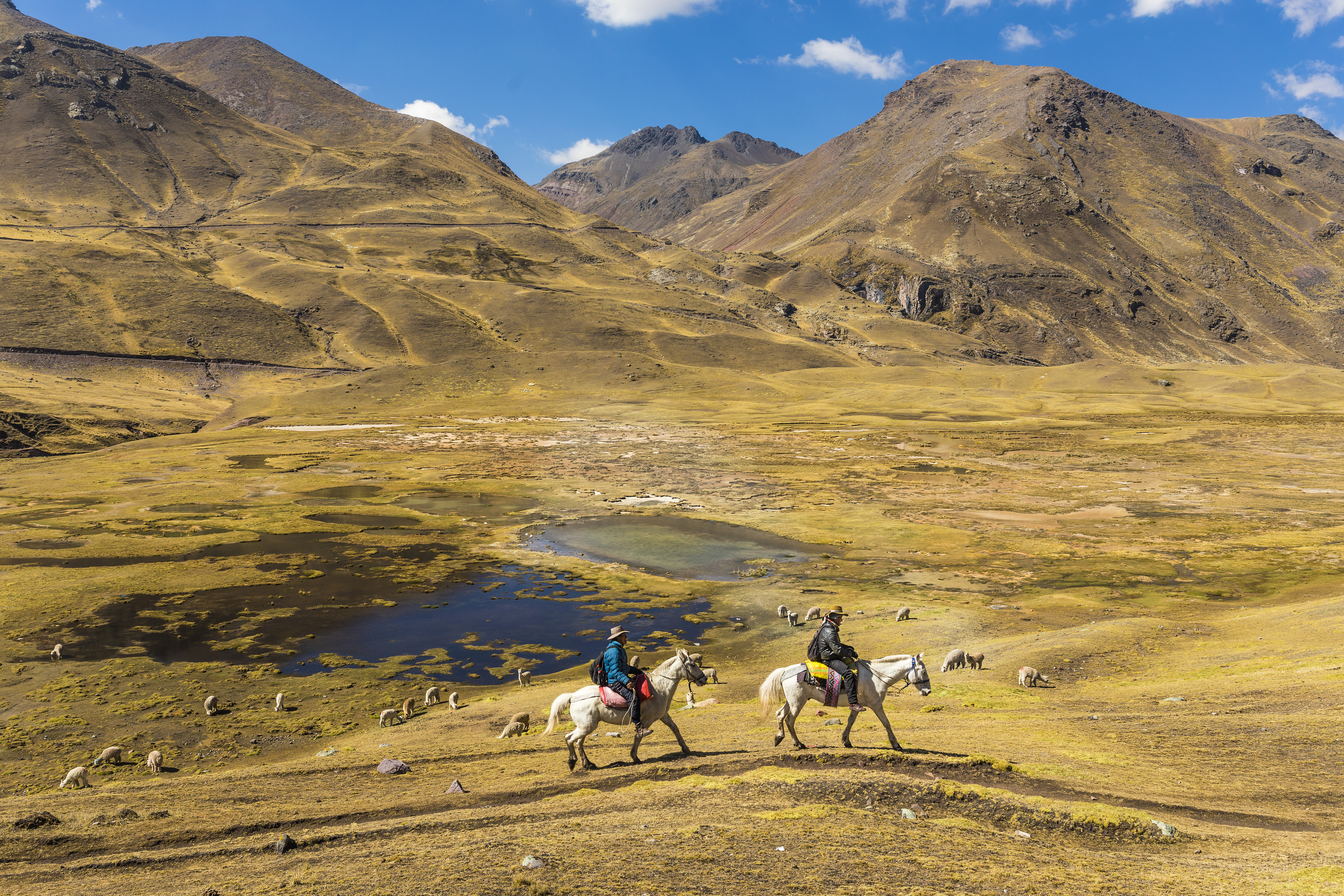 Horseback riders cross highland grass beside a winding river and small lakes, surrounded by rugged brown mountains all around them.