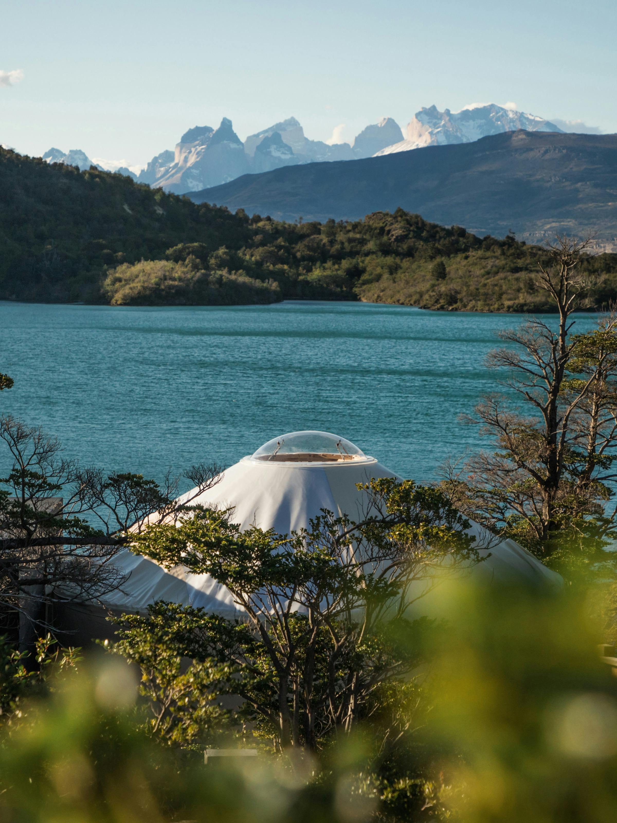White dome tent sits beside a blue lake, with snowy mountains rising beyond green shrubs in foreground nearby under clouds.