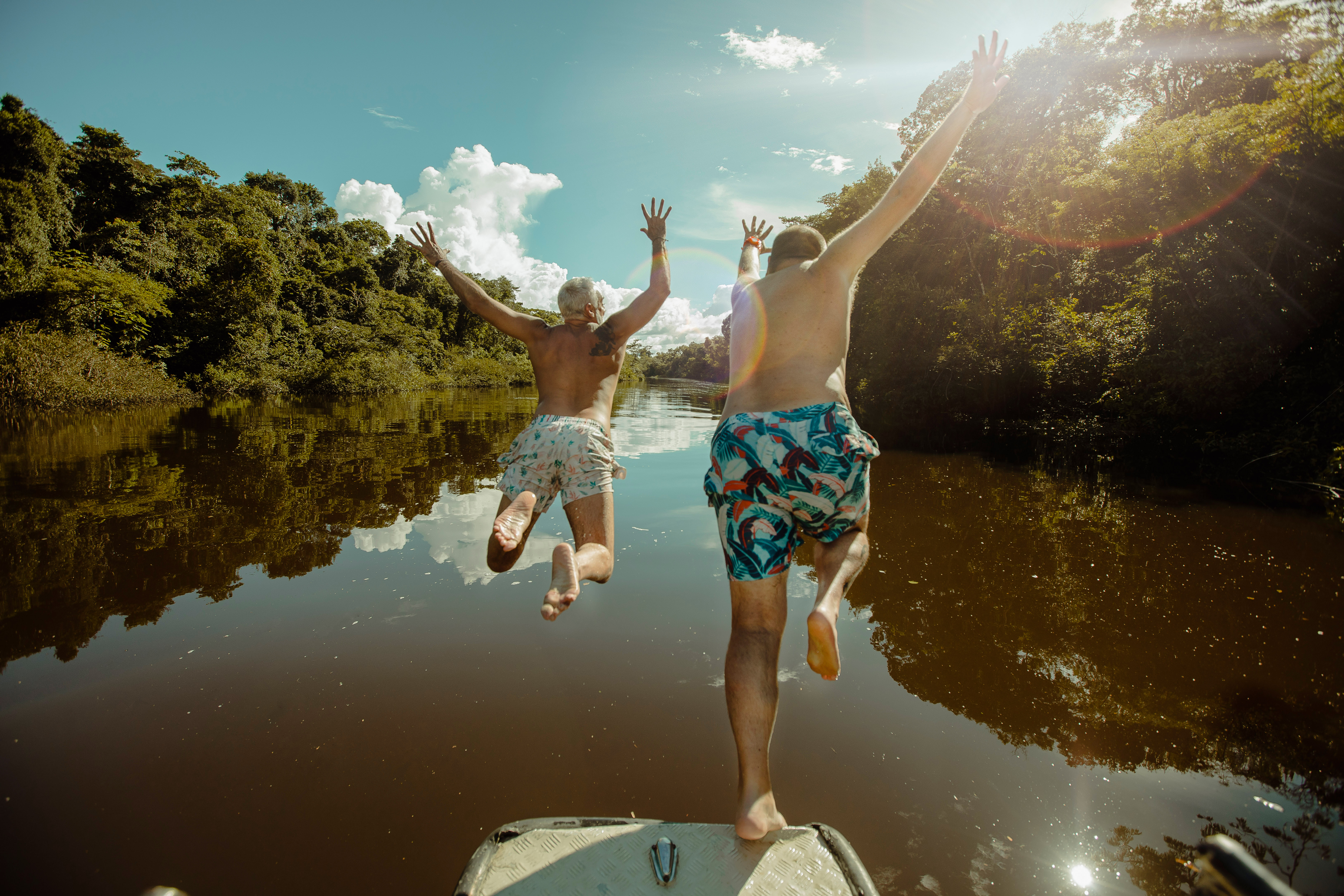 Two people leap from a boat into a calm river, splashing beside jungle banks and sunlit reflections below them.