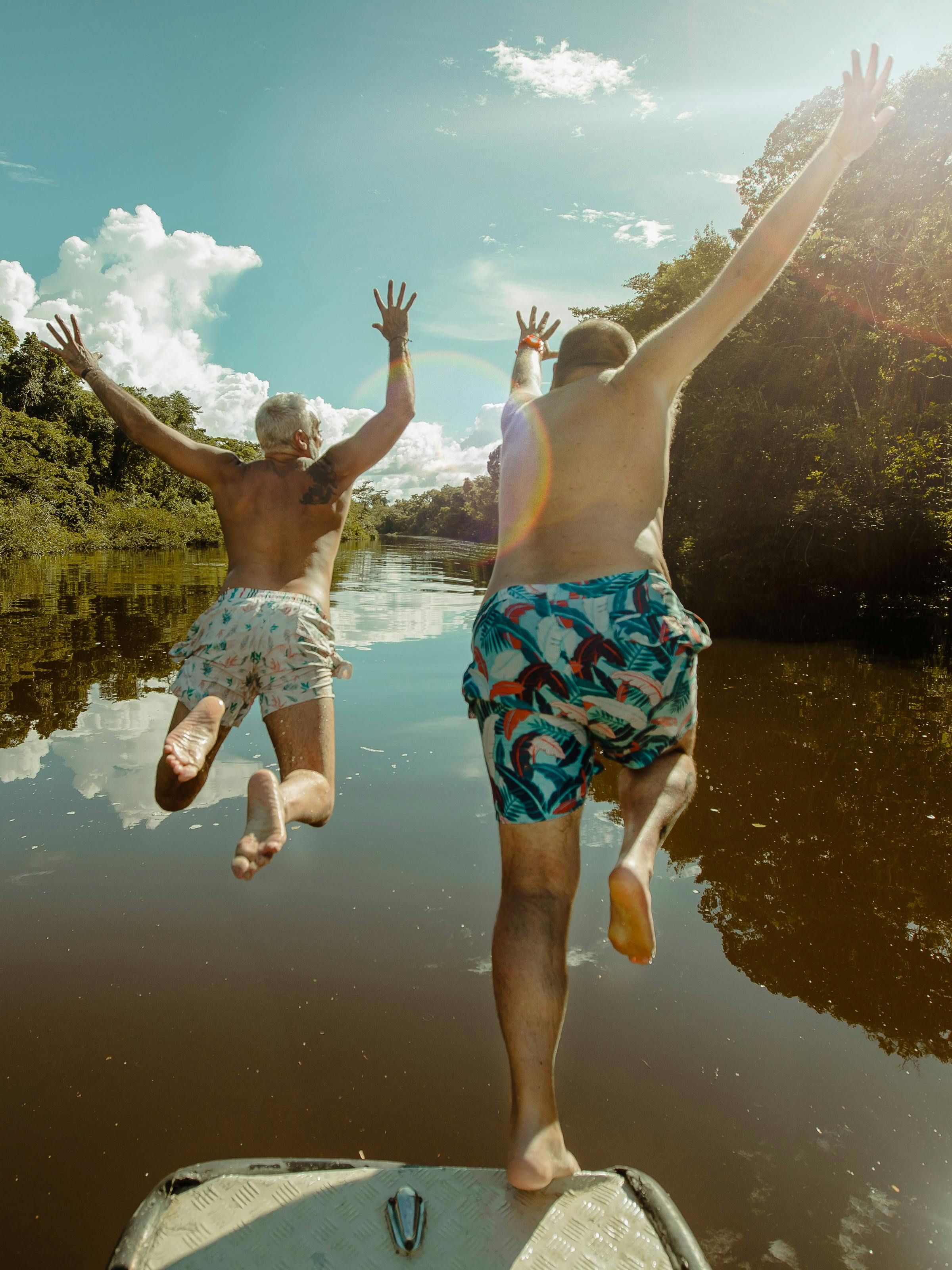 Two people leap from a boat into a calm river, splashing beside jungle banks and sunlit reflections below them.
