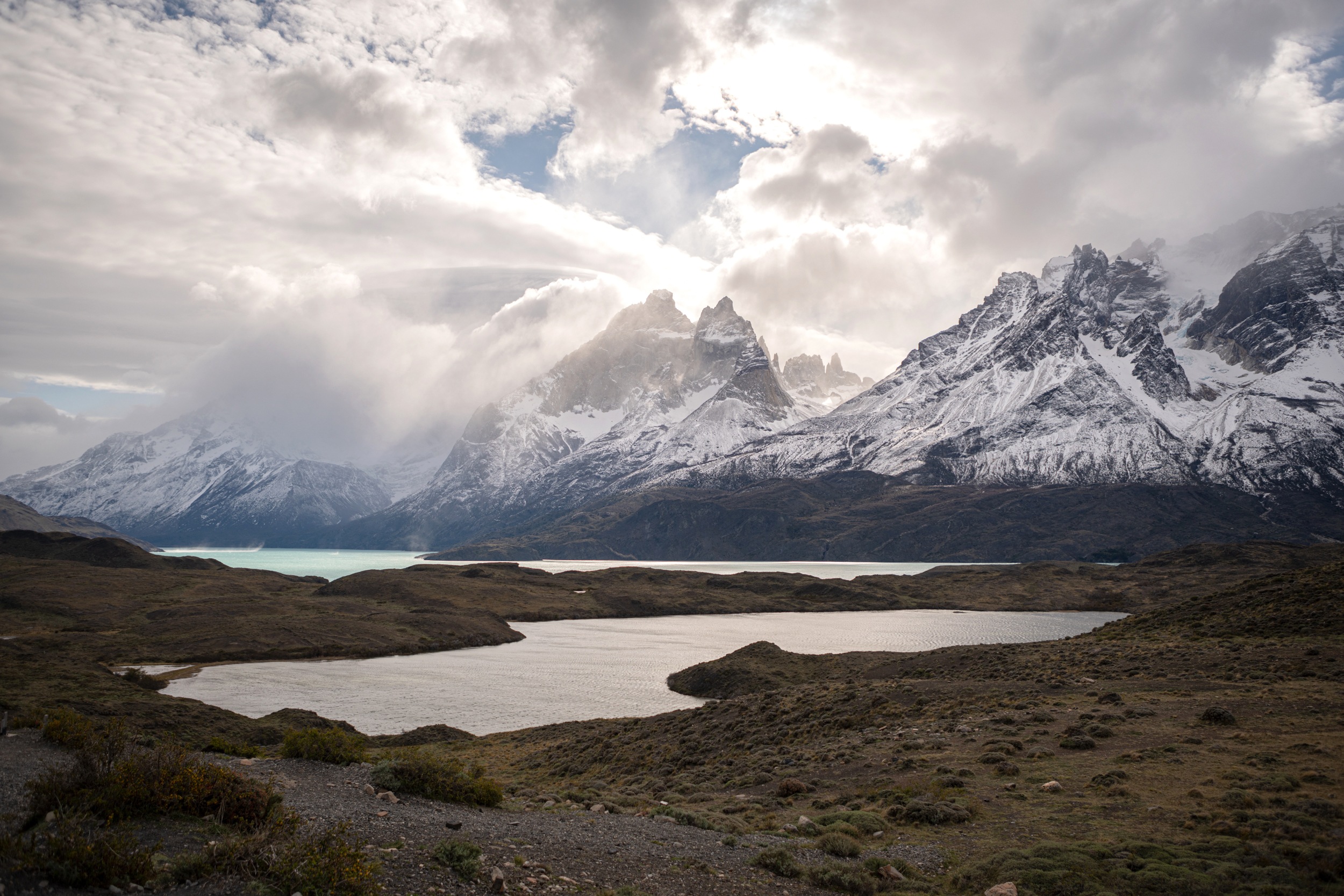 Wide mountain landscape with a dark lake below snow-dusted peaks, under heavy clouds and soft filtered light across the valley.