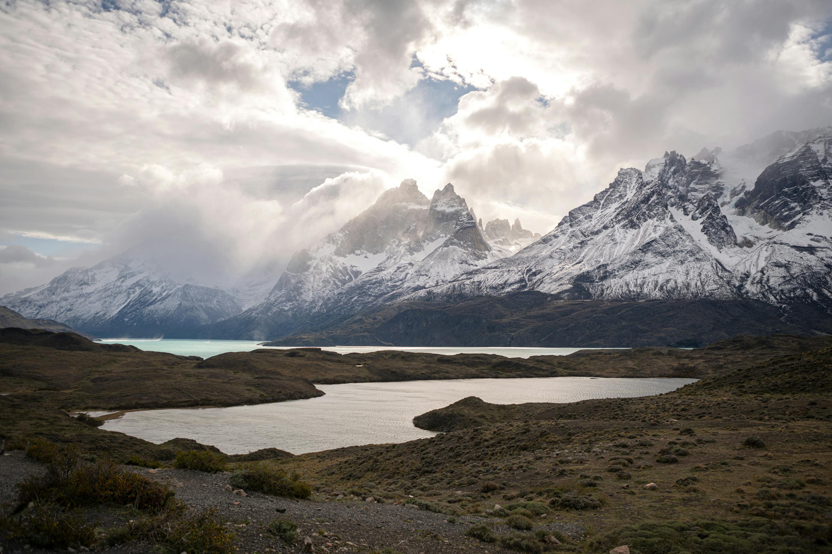 Wide mountain landscape with a dark lake below snow-dusted peaks, under heavy clouds and soft filtered light across the valley.