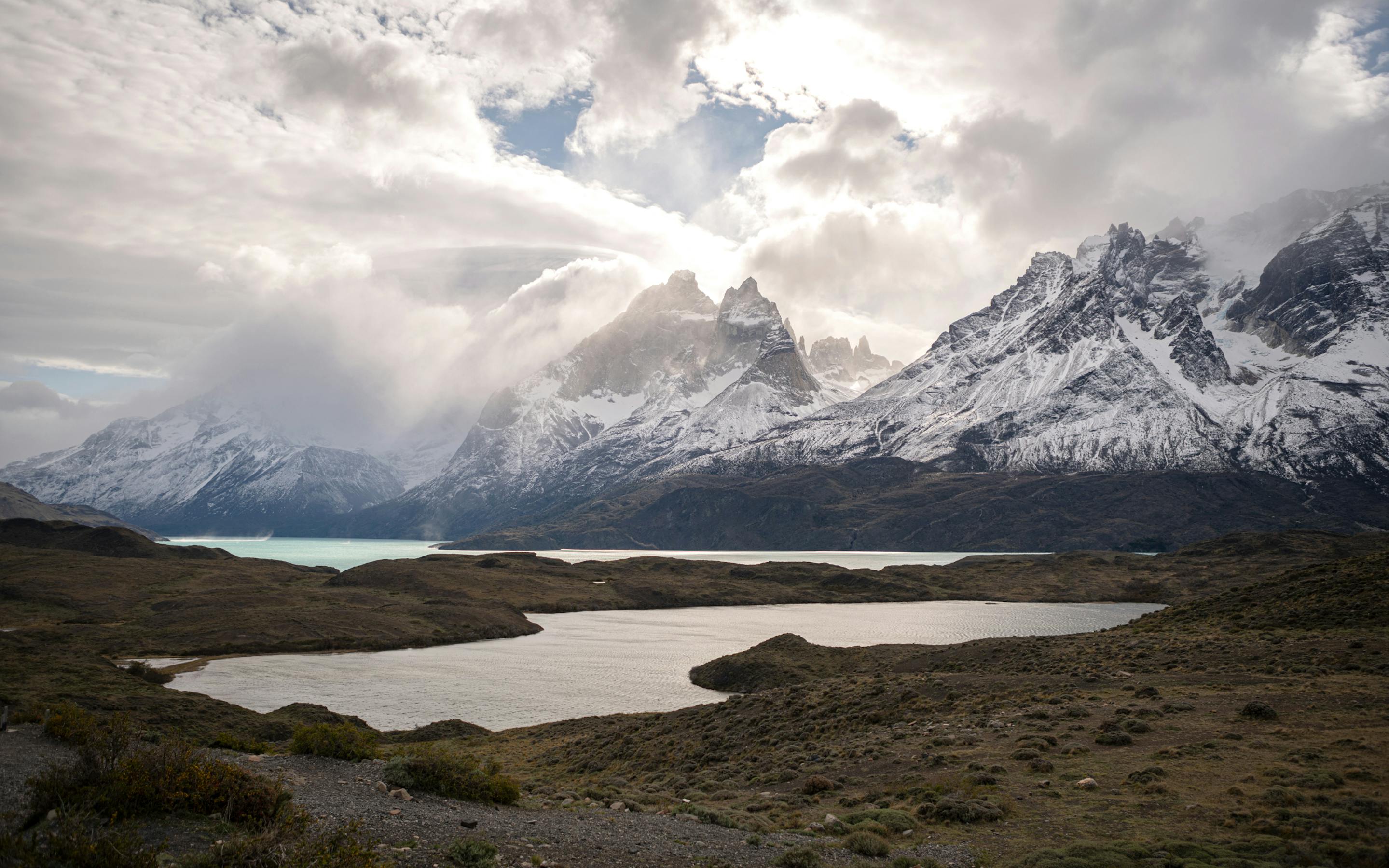 Wide mountain landscape with a dark lake below snow-dusted peaks, under heavy clouds and soft filtered light across the valley.