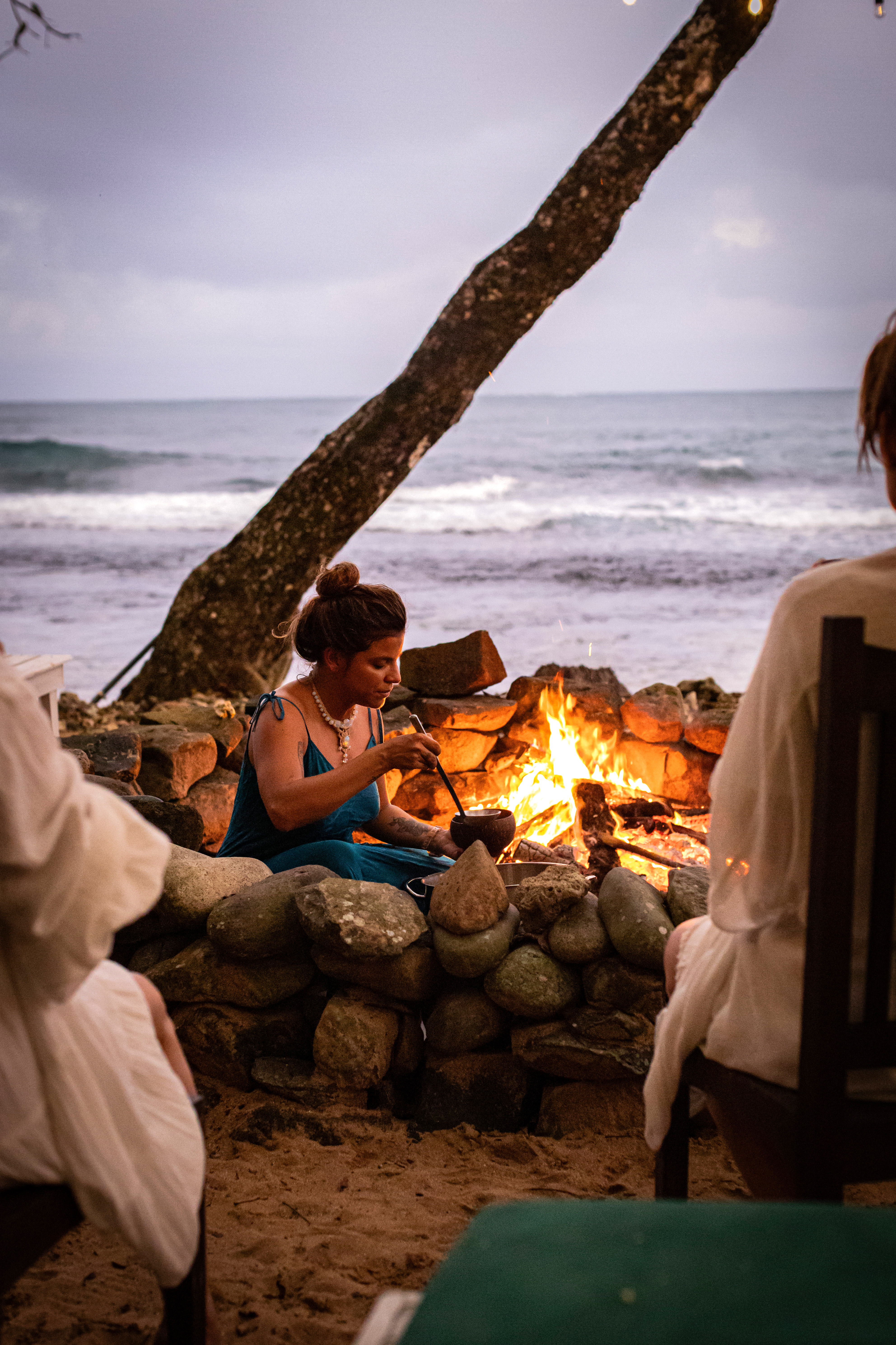 People gather around a small bonfire on sandy beach at dusk, with waves, clouds, and a leaning palm behind them.