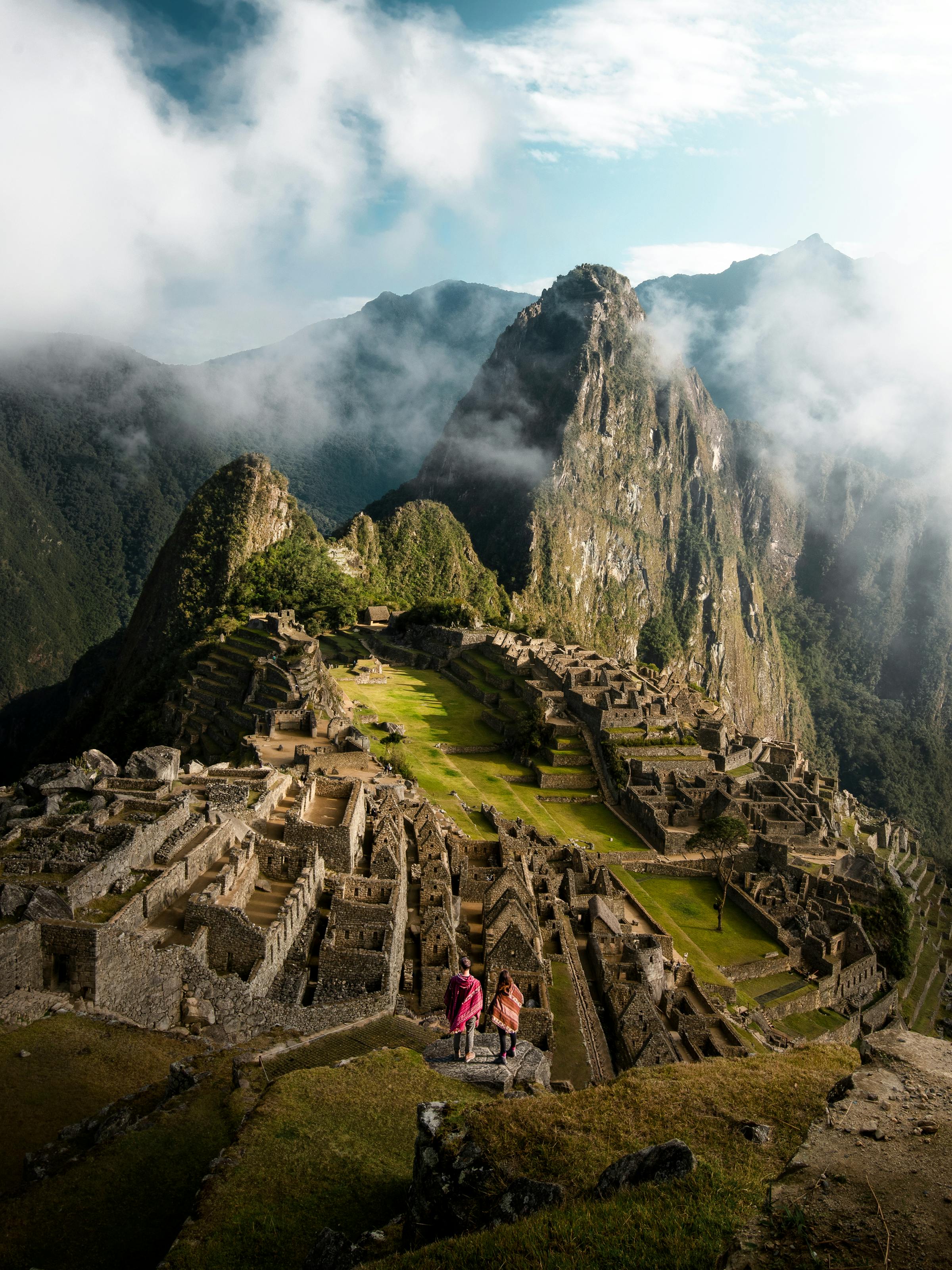 Ancient stone terraces and ruins sit on a steep green ridge, with dramatic peaks and low clouds surrounding them nearby.