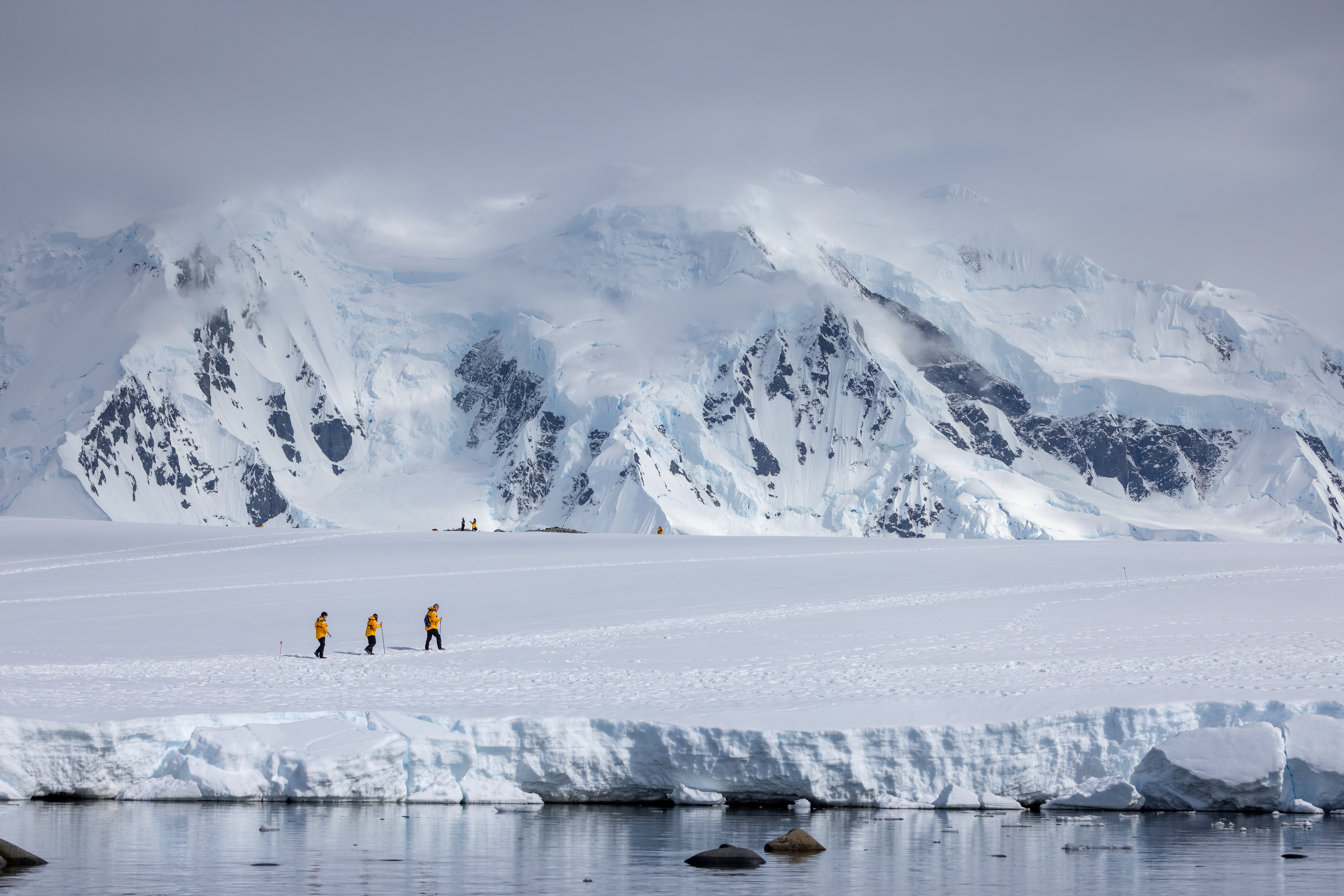 Tiny figures walk across a vast snowy plain beneath steep white mountains, with blowing clouds and bright light above them.