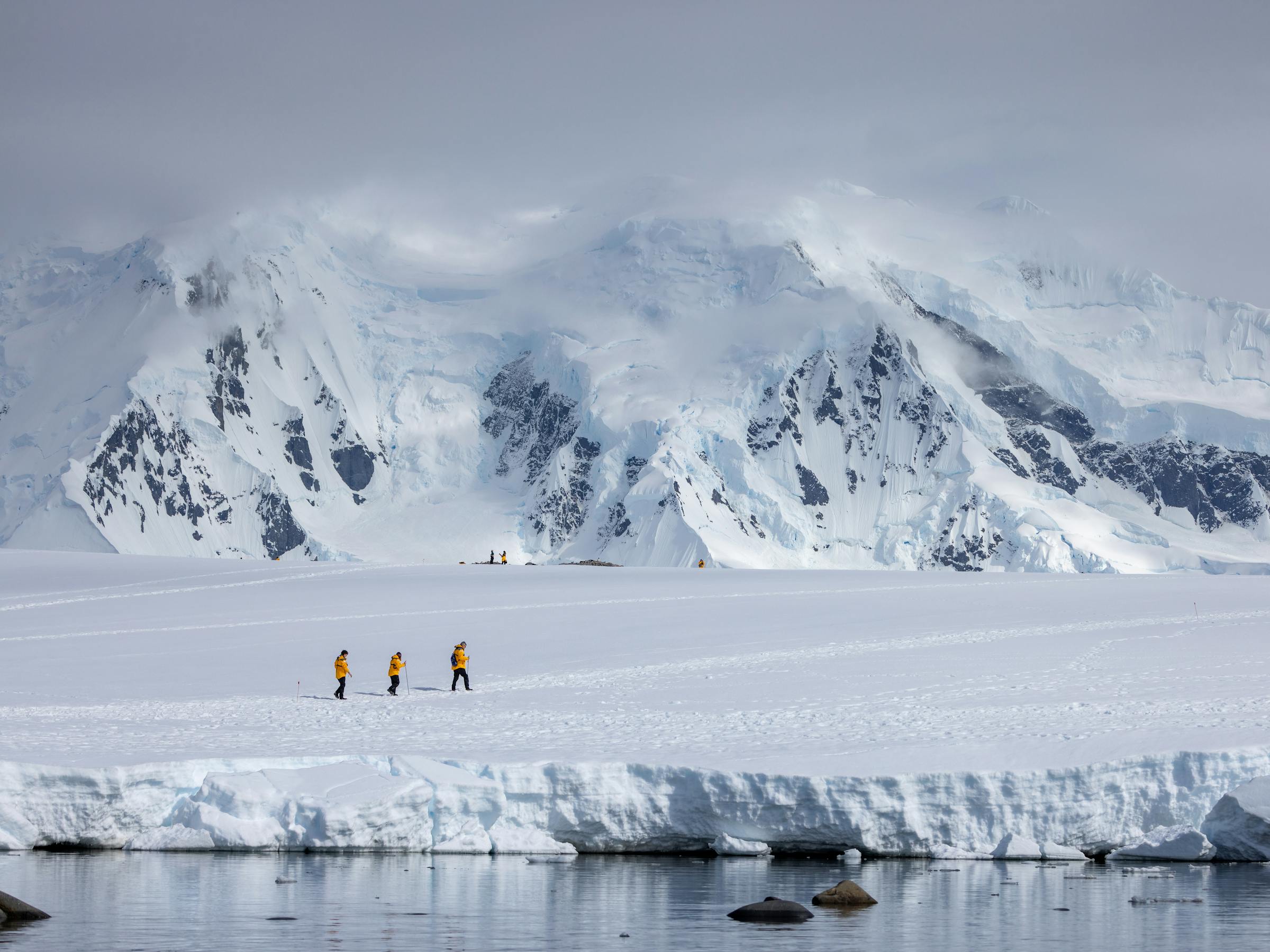 Tiny figures walk across a vast snowy plain beneath steep white mountains, with blowing clouds and bright light above them.