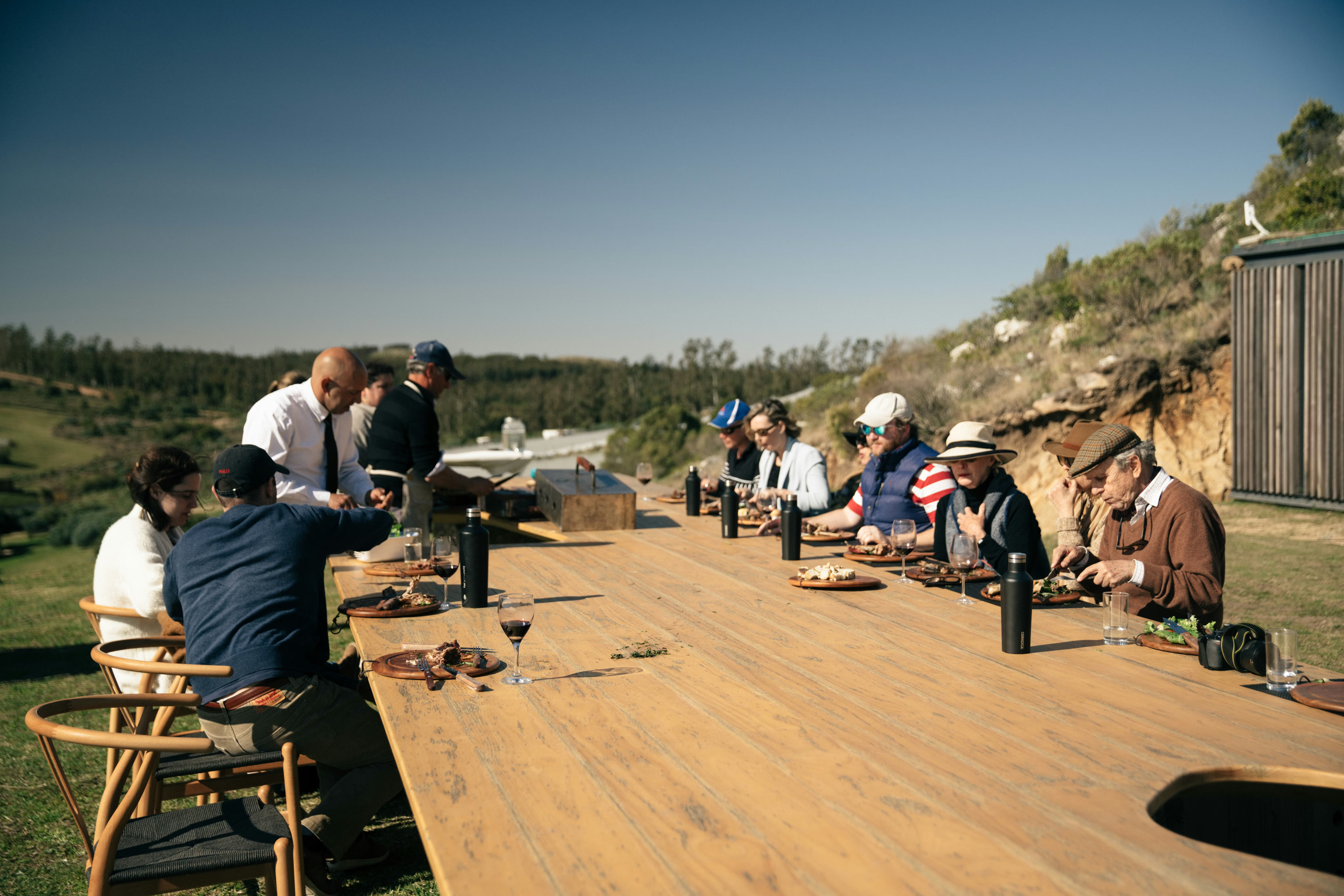 People sit at long wooden tables on an outdoor terrace overlooking rolling hills, with clear sky and distant ridges.