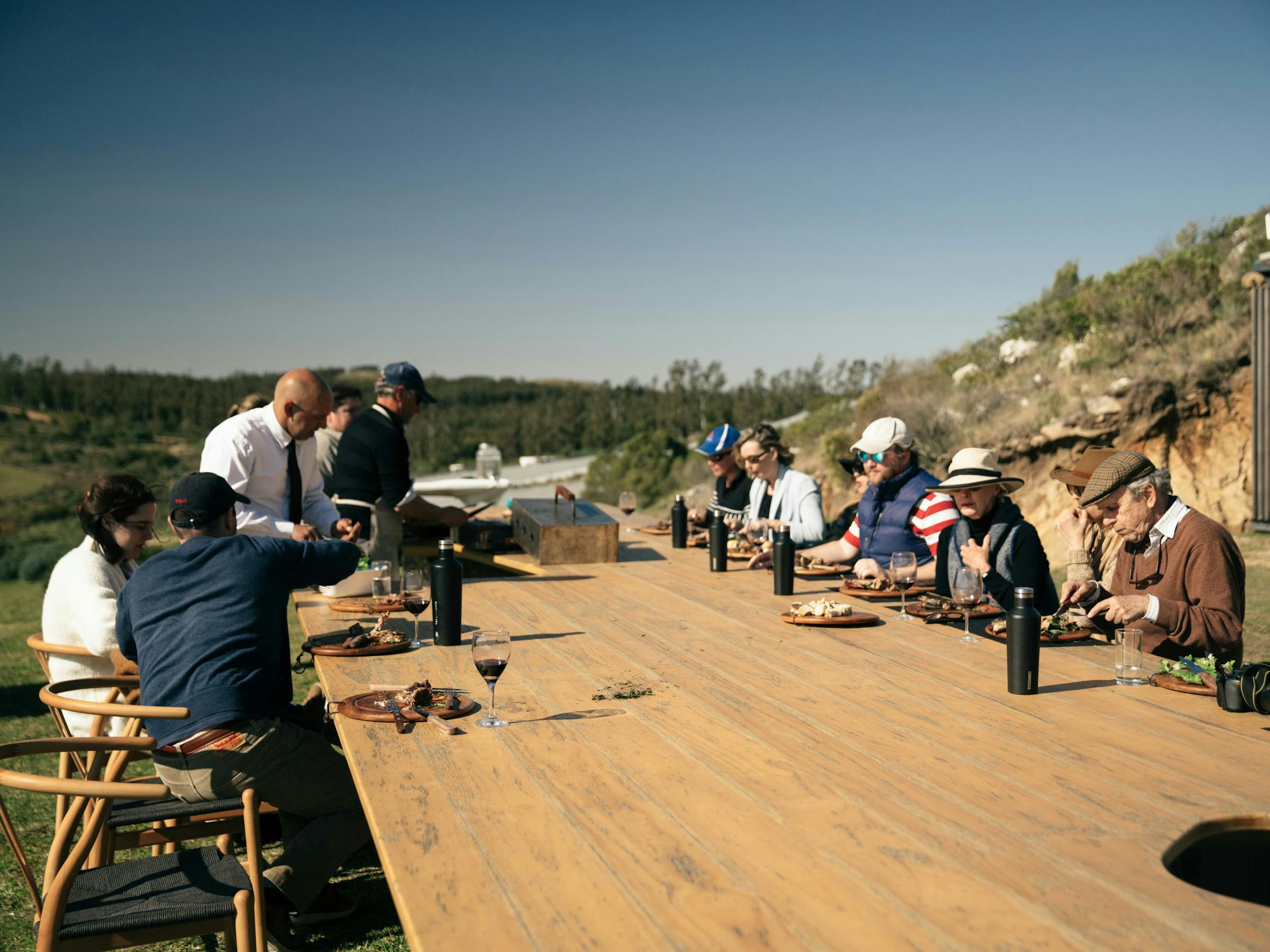 People sit at long wooden tables on an outdoor terrace overlooking rolling hills, with clear sky and distant ridges.