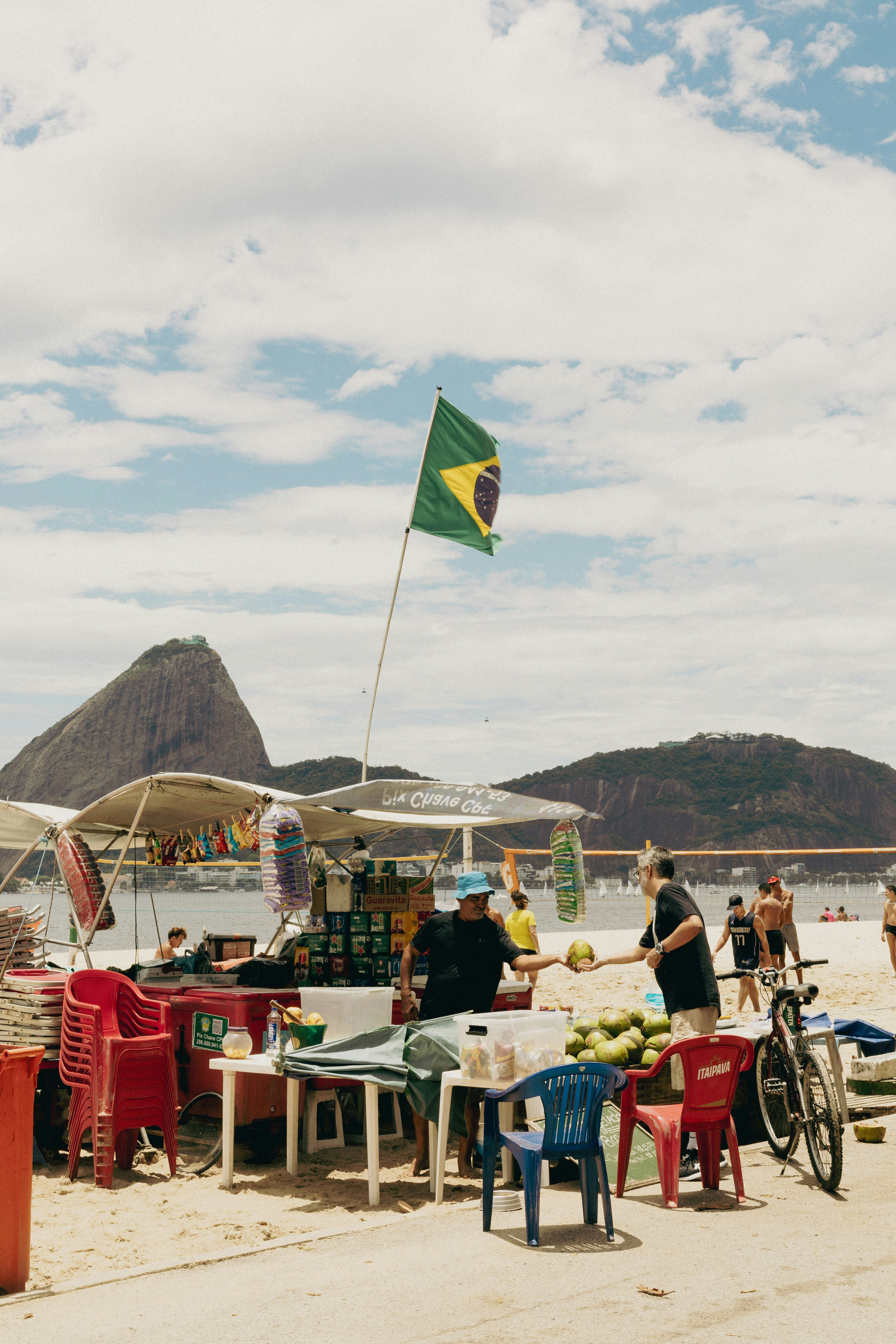 Outdoor market scene with Brazilian flag and people seated at tables, with mountains and blue sky in distance behind them.