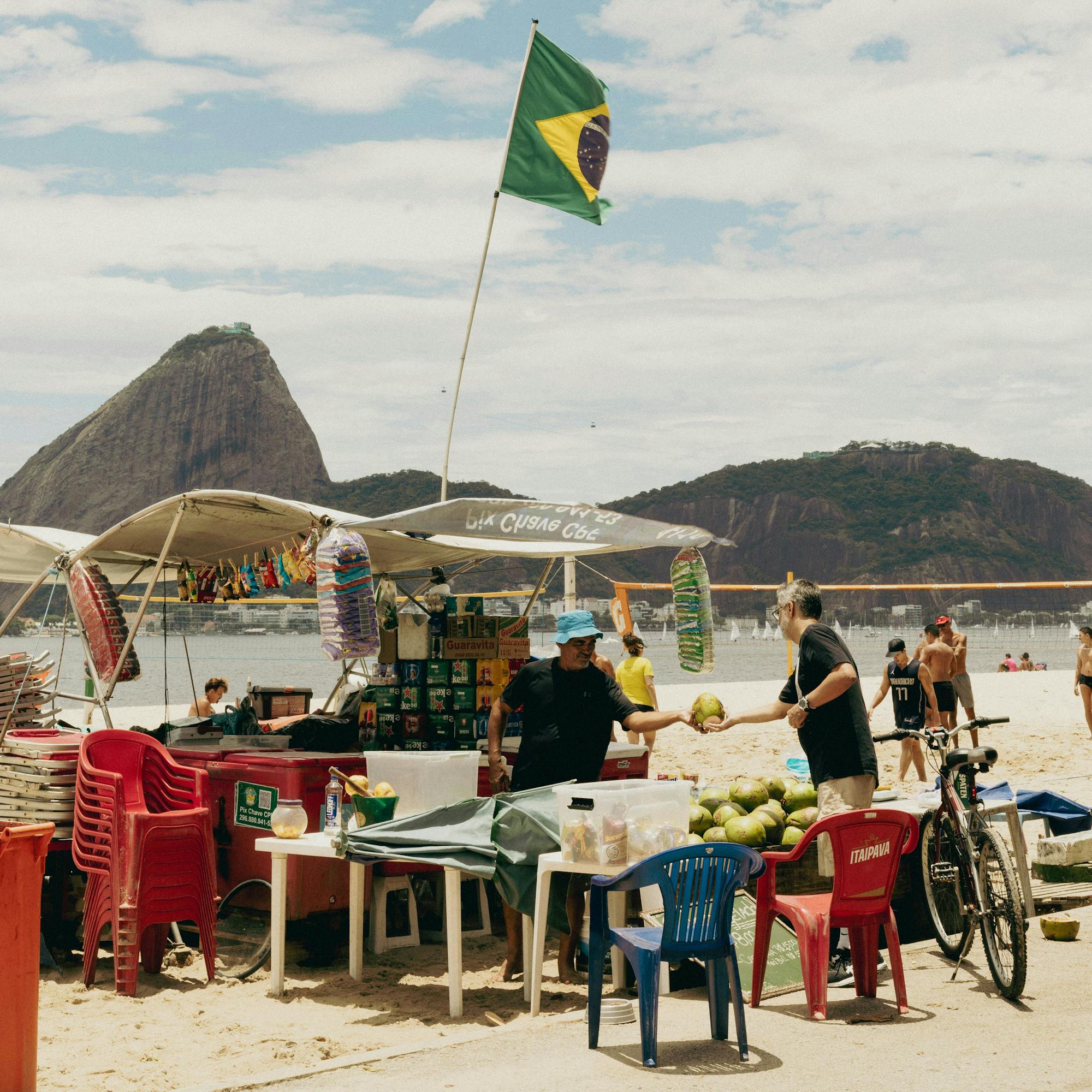 Outdoor market scene with Brazilian flag and people seated at tables, with mountains and blue sky in distance behind them.