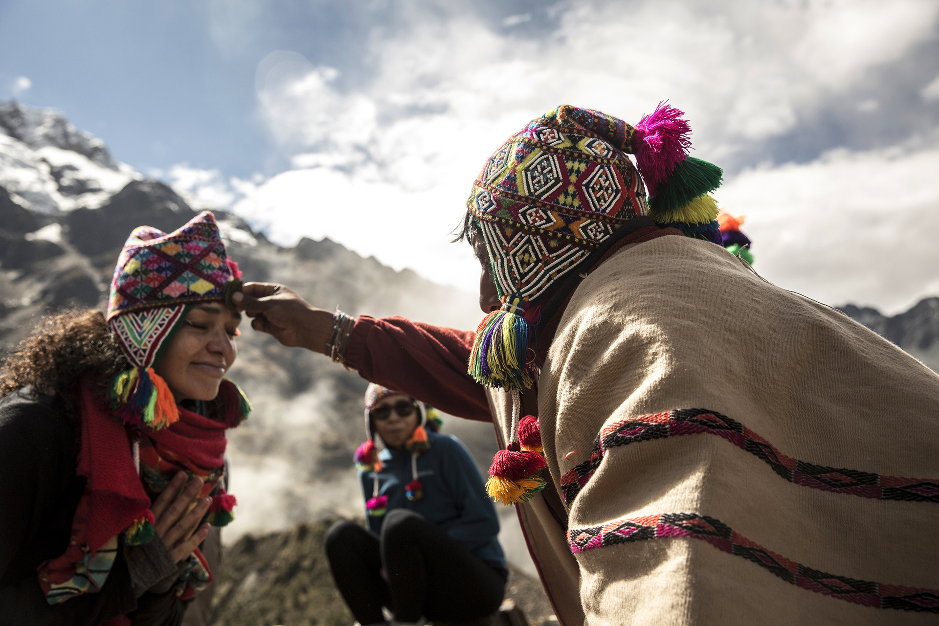 Two people in traditional woven clothing speak on a rocky overlook, with snowcapped peaks and clouds behind them outside.