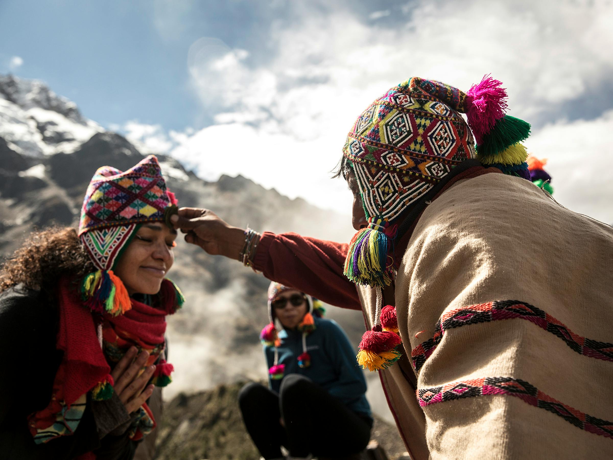 Two people in traditional woven clothing speak on a rocky overlook, with snowcapped peaks and clouds behind them outside.