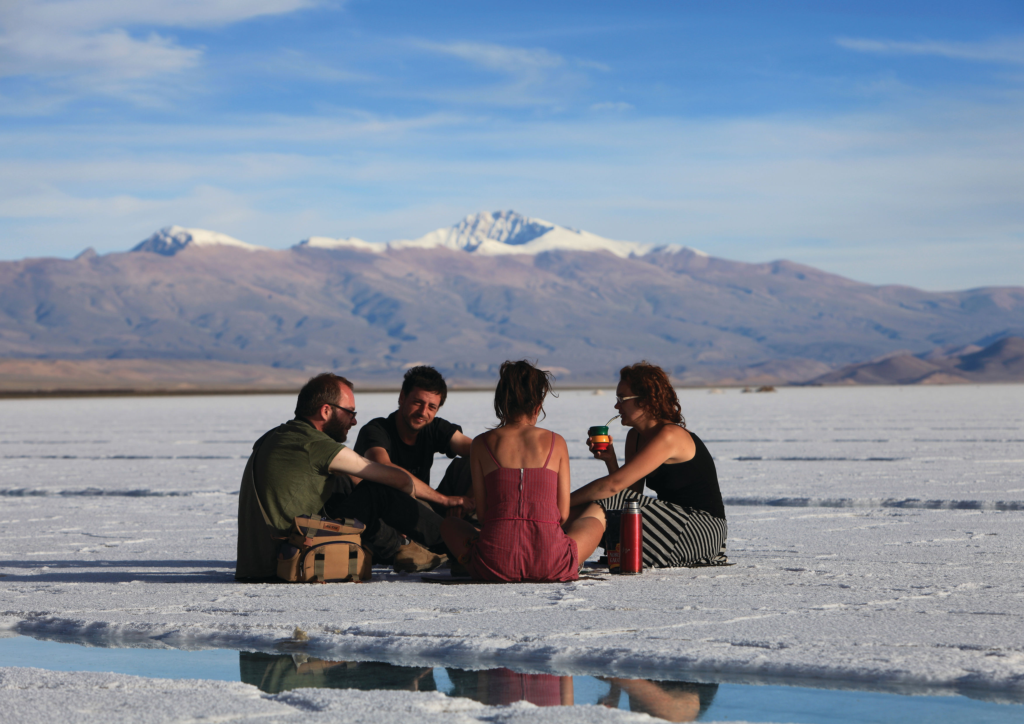 A group of people sit on a white salt flat with distant mountains, while blue sky stretches across the horizon behind them.