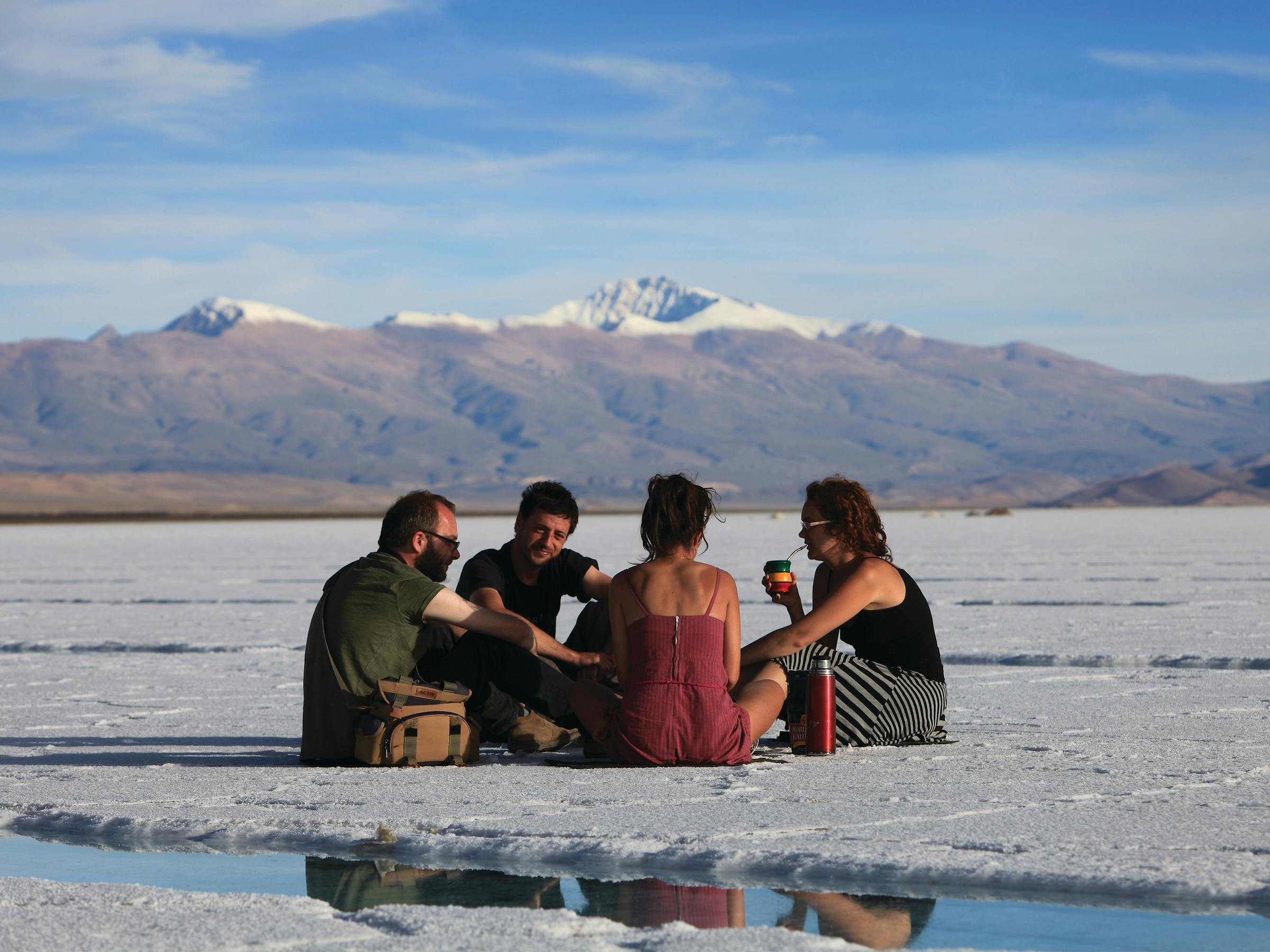 A group of people sit on a white salt flat with distant mountains, while blue sky stretches across the horizon behind them.